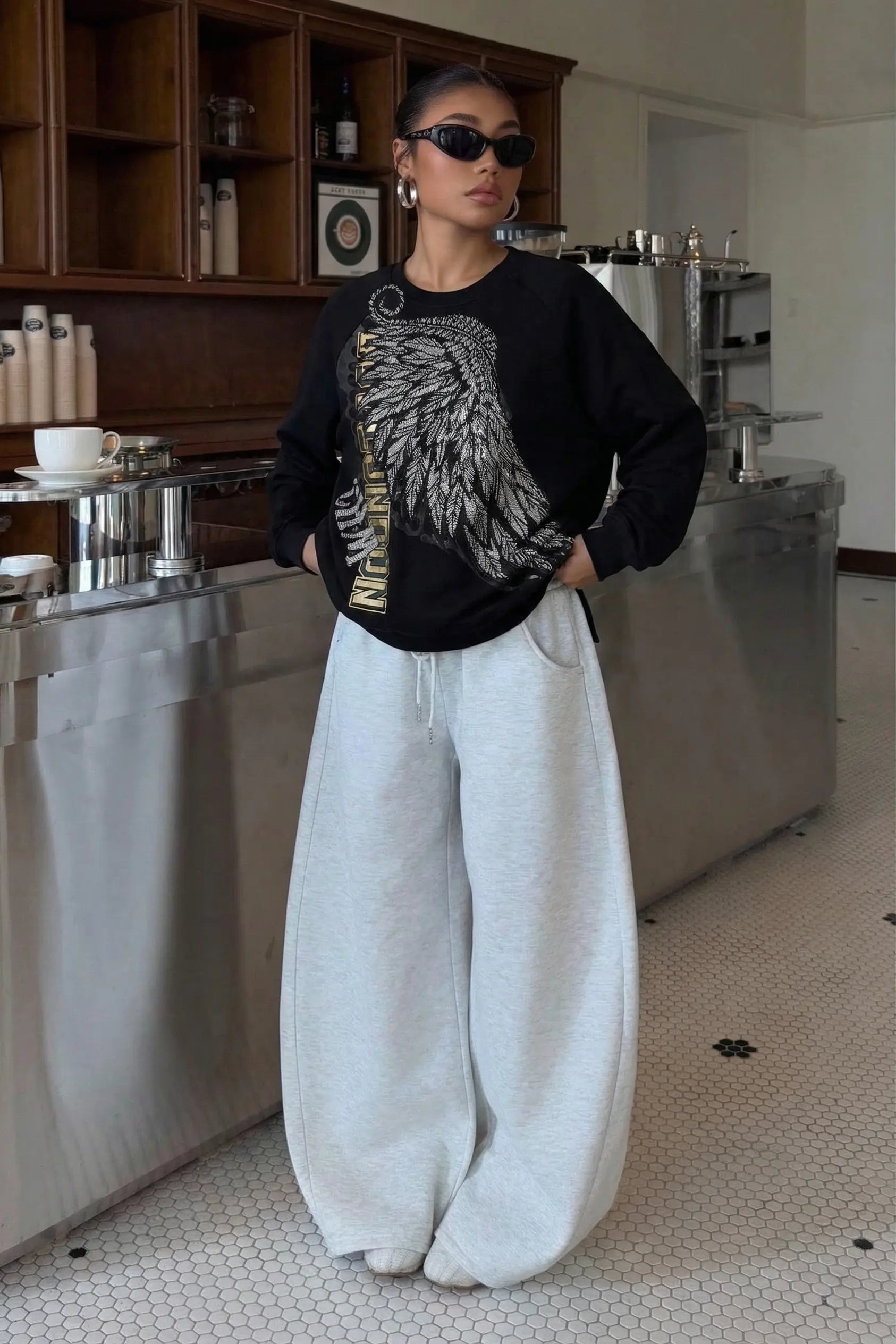 Person wearing a black sweater with a graphic design and white pants in a kitchen setting, Rhinestone Wing Embroidered Sweatshirt