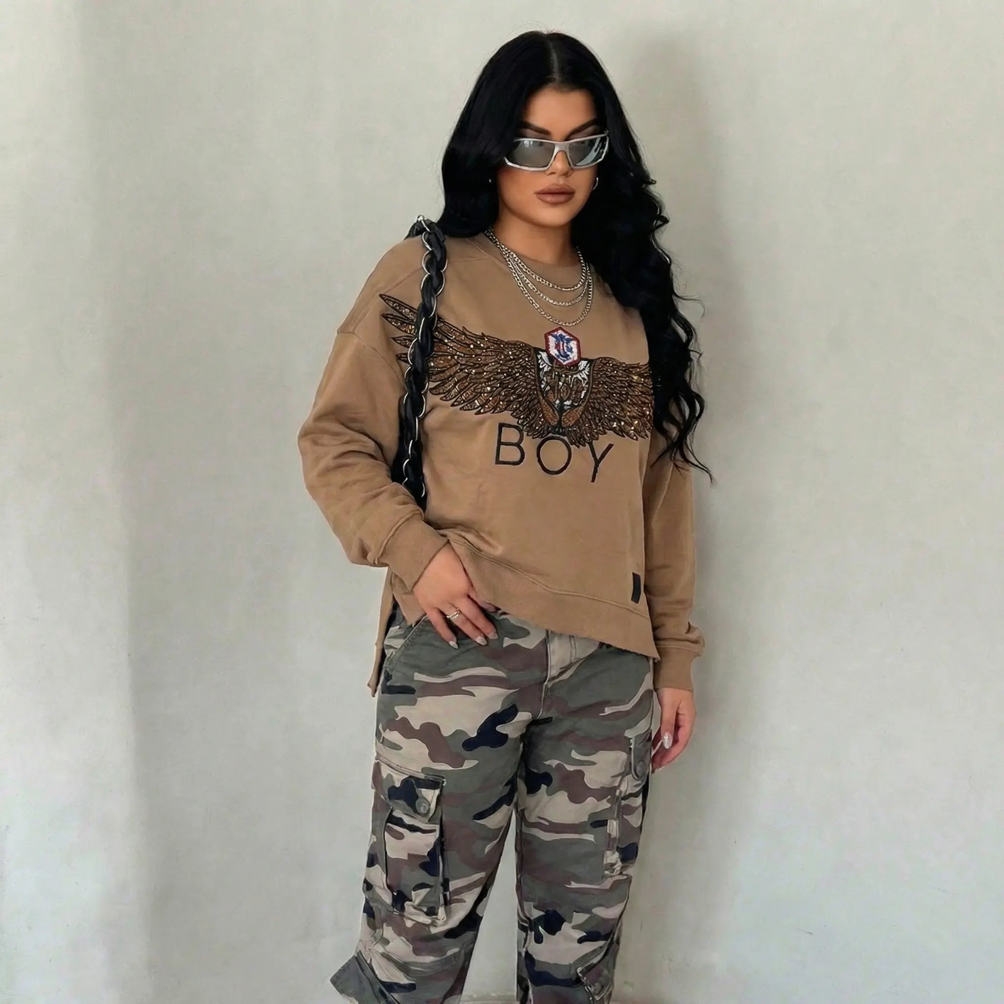 Person wearing a beige sweatshirt with an eagle design and 'BOY' text, paired with camouflage pants against a plain background. Rhinestone Wing Logo Sweatshirt