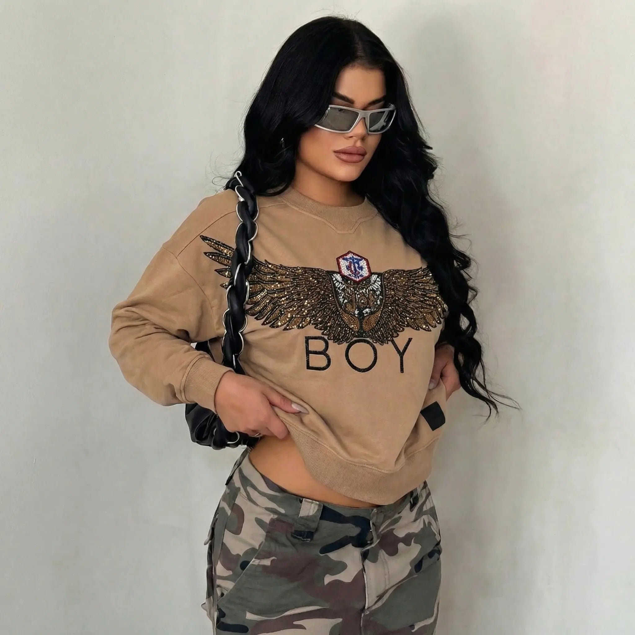 Person wearing a beige sweatshirt with 'BOY' and camouflage pants against a plain background, Rhinestone Wing Logo Sweatshirt