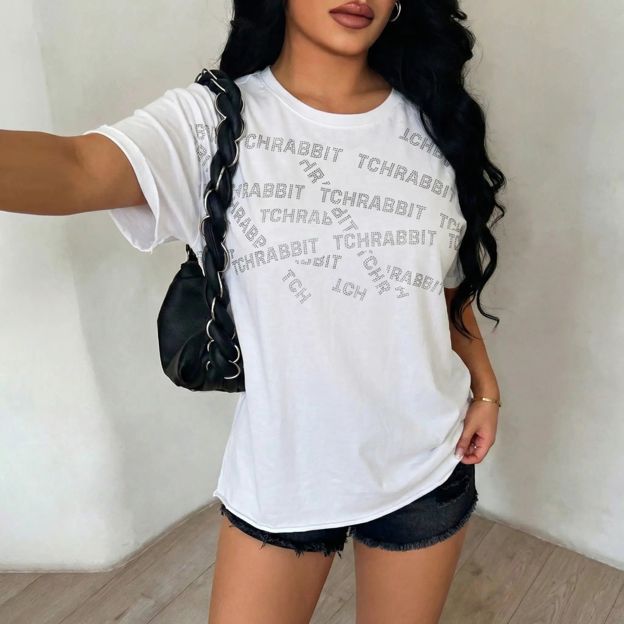 Person wearing a white t-shirt with text and black shorts, holding a black bag. TCH All-Over TCHRABBIT Logo T-Shirt