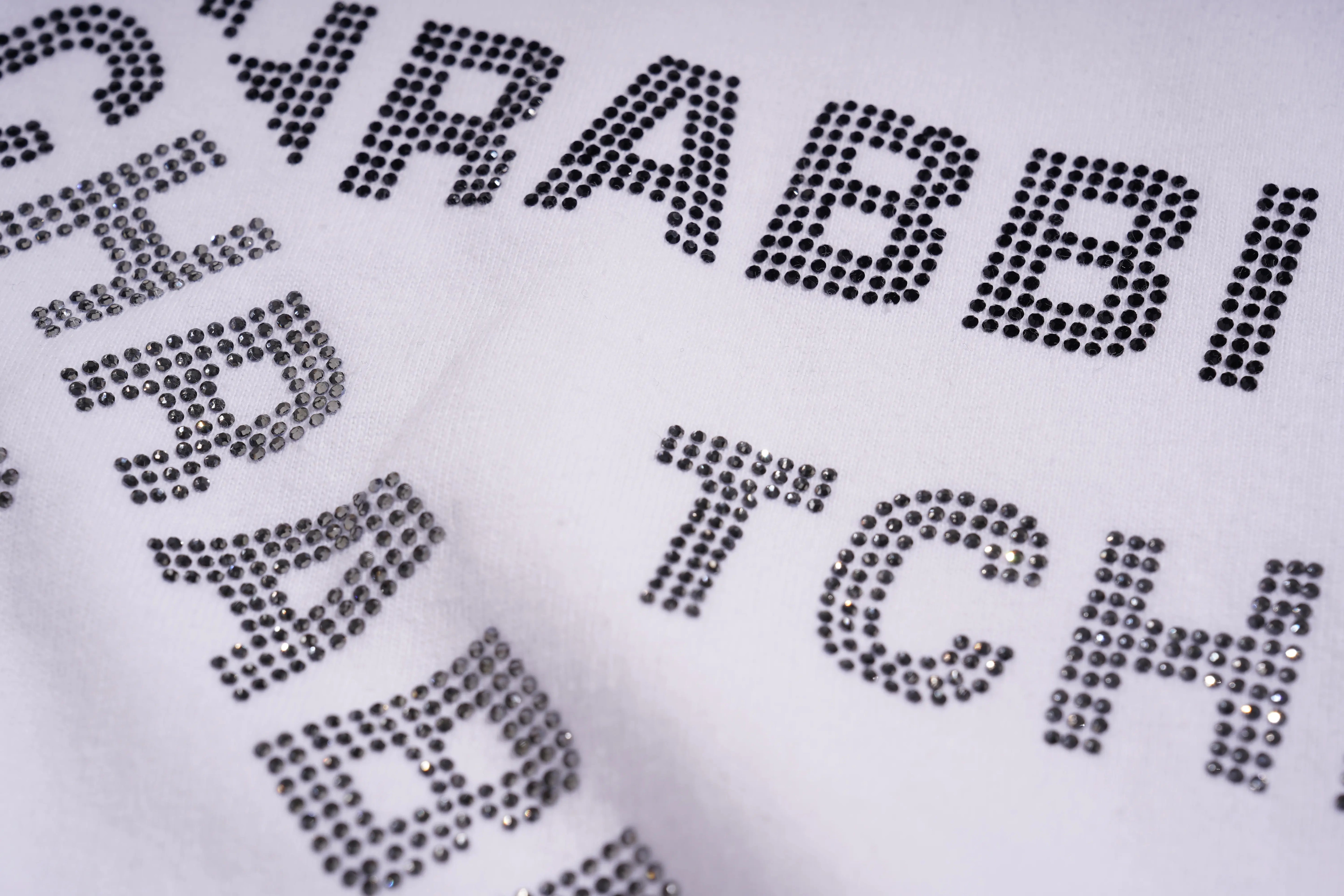 Textured fabric with 'RABBI' and 'THE' printed in rhinestone-like black stones on a white background, TCH All-Over TCHRABBIT Logo T-Shirt