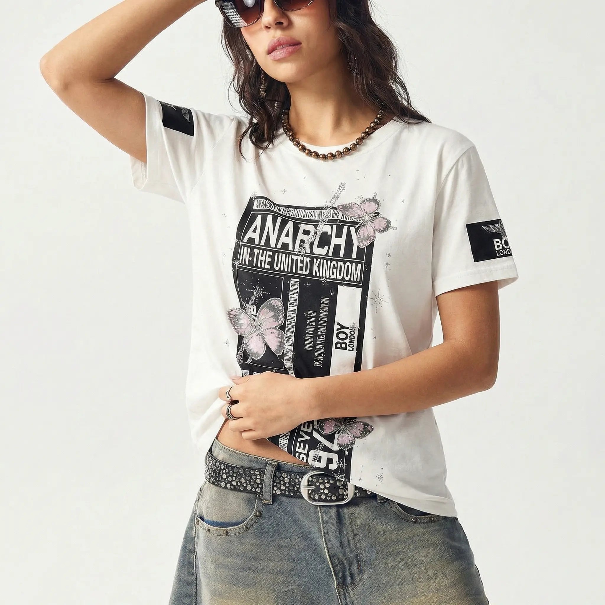 Person wearing a white t-shirt with black graphics and text, denim jeans, and sunglasses on a light gray background, TCH Anarchy Graphic Rhinestone T-Shirt