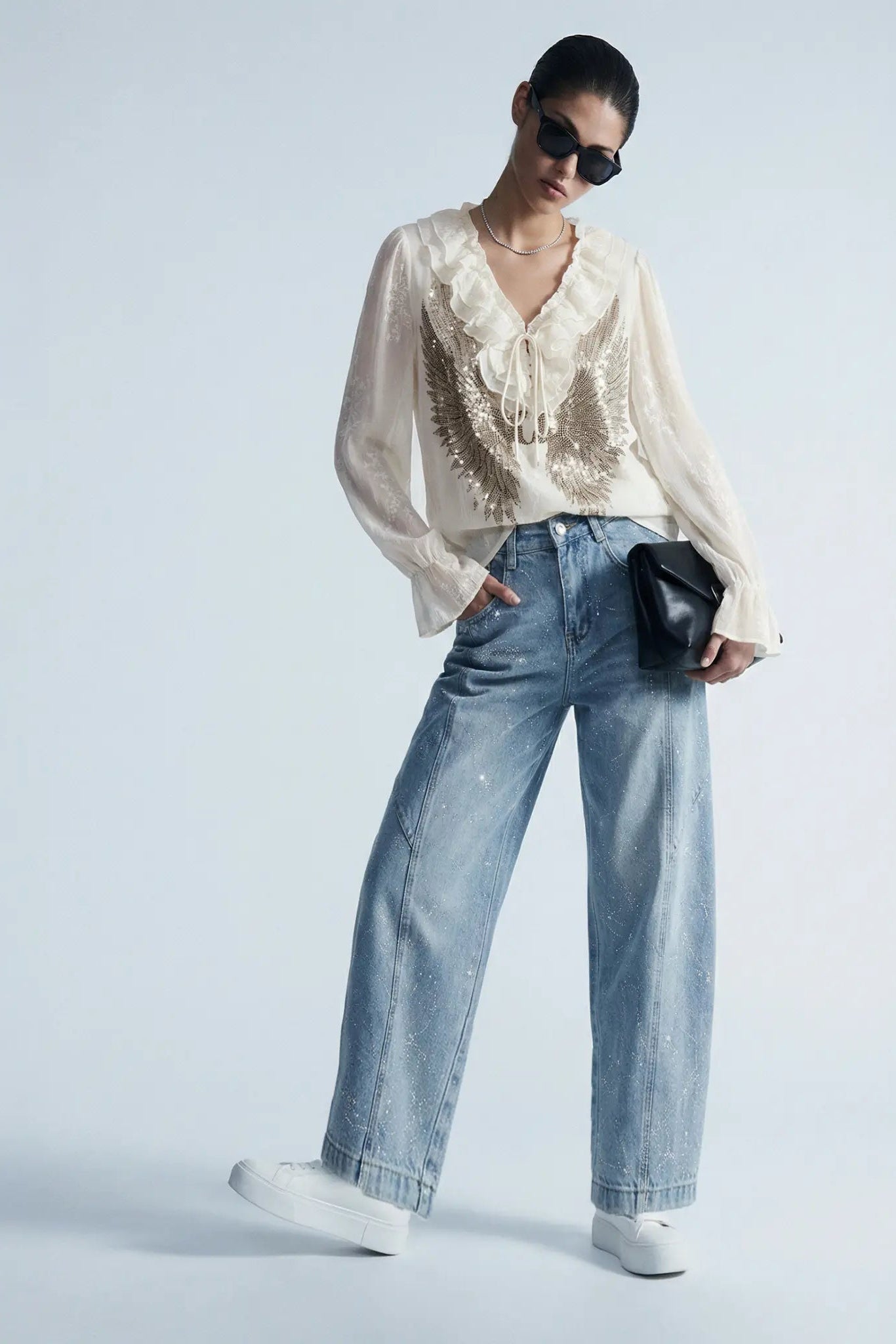 Person wearing a white blouse with lace details and blue jeans on a light gray background, TCH Angel Wings Rhinestone Ruffle Blouse