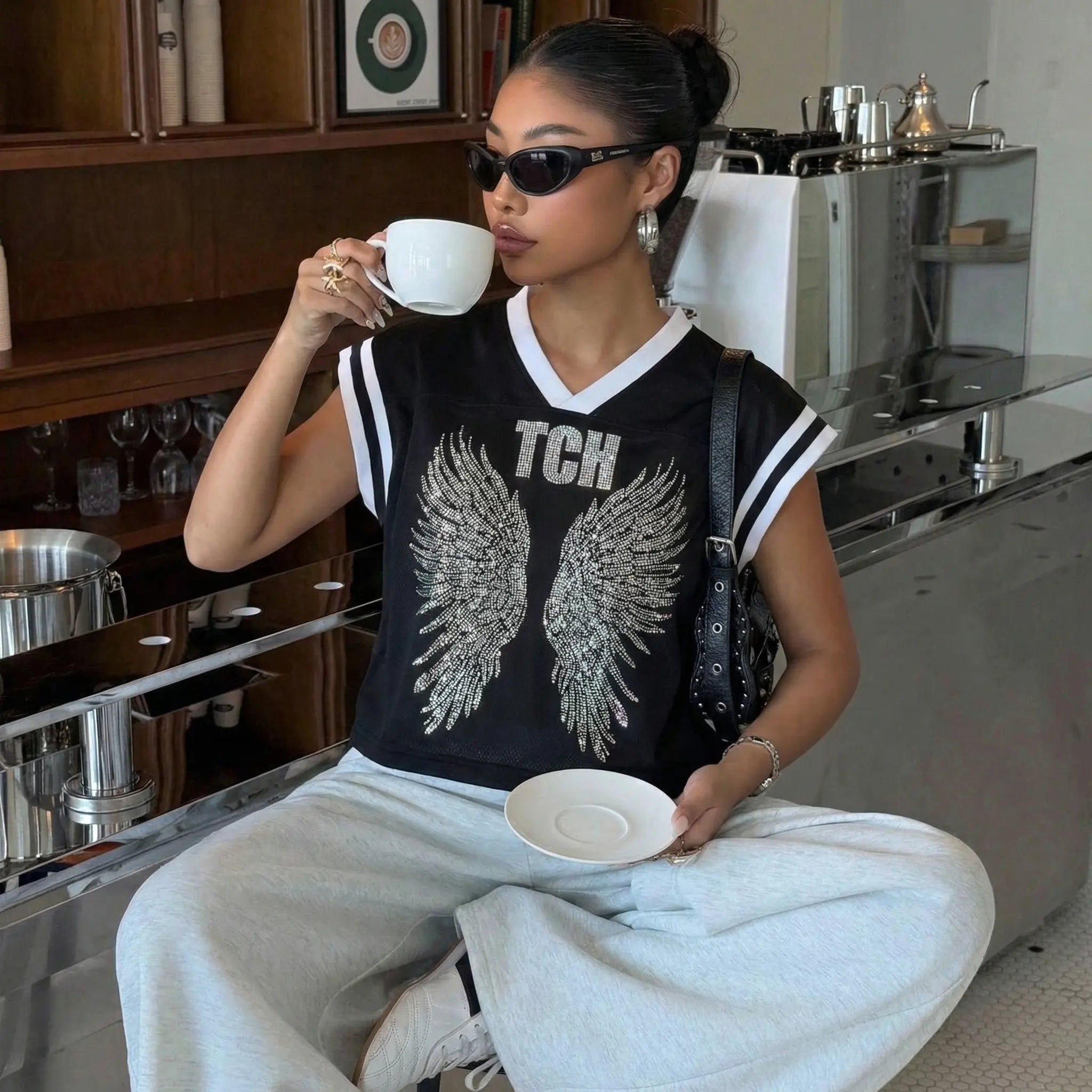 Woman sitting in a kitchen holding a cup and saucer, wearing a black 'TCH' shirt with wings. TCH Angel Wings Rhinestone V-Neck Tee