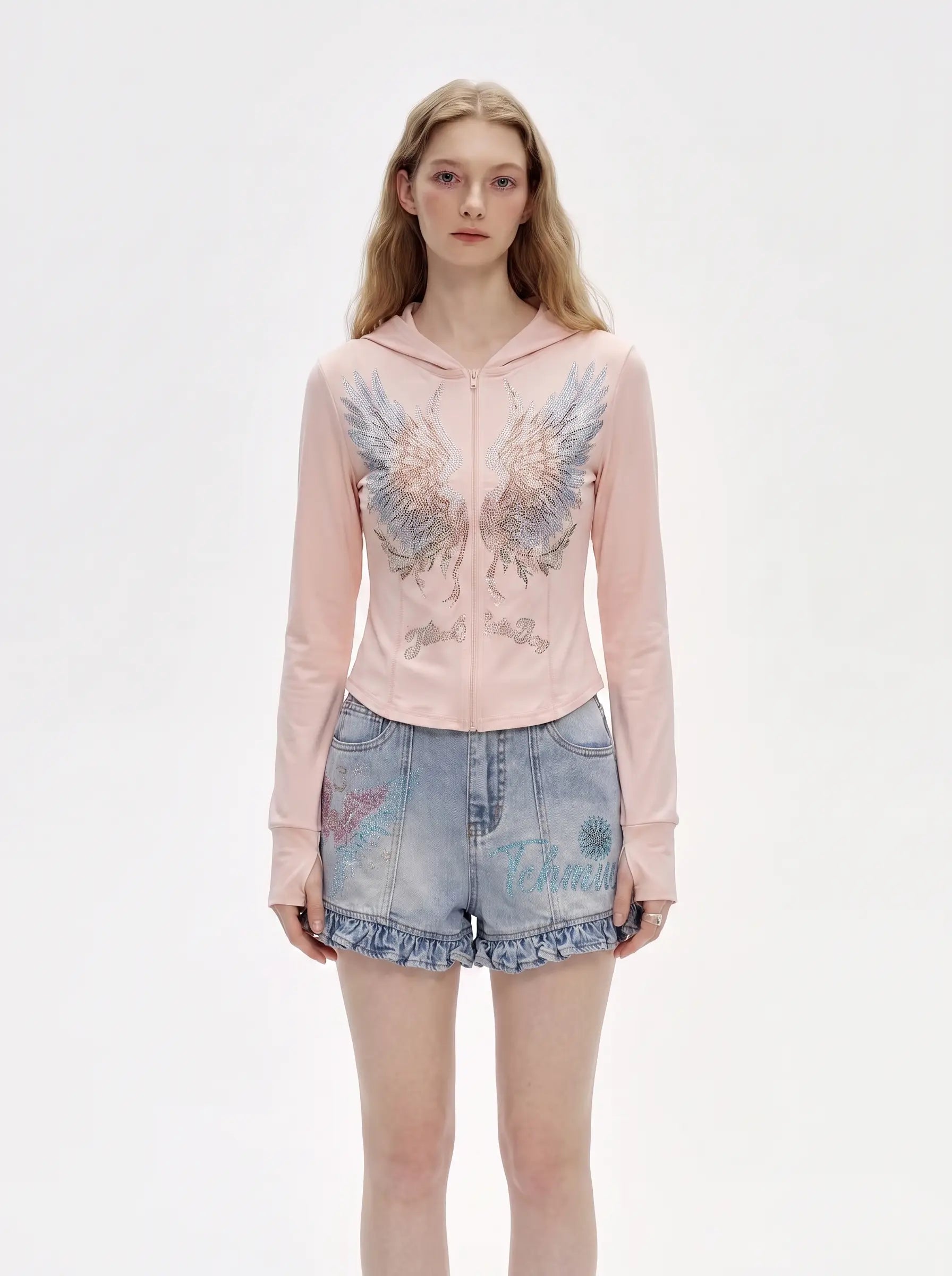 A model wearing a light pink zip-up hoodie with a large angel wing graphic on the front, paired with light-wash denim shorts, against a plain white studio background. TCH Angel Wings Rhinestone Zip-Up Hoodie