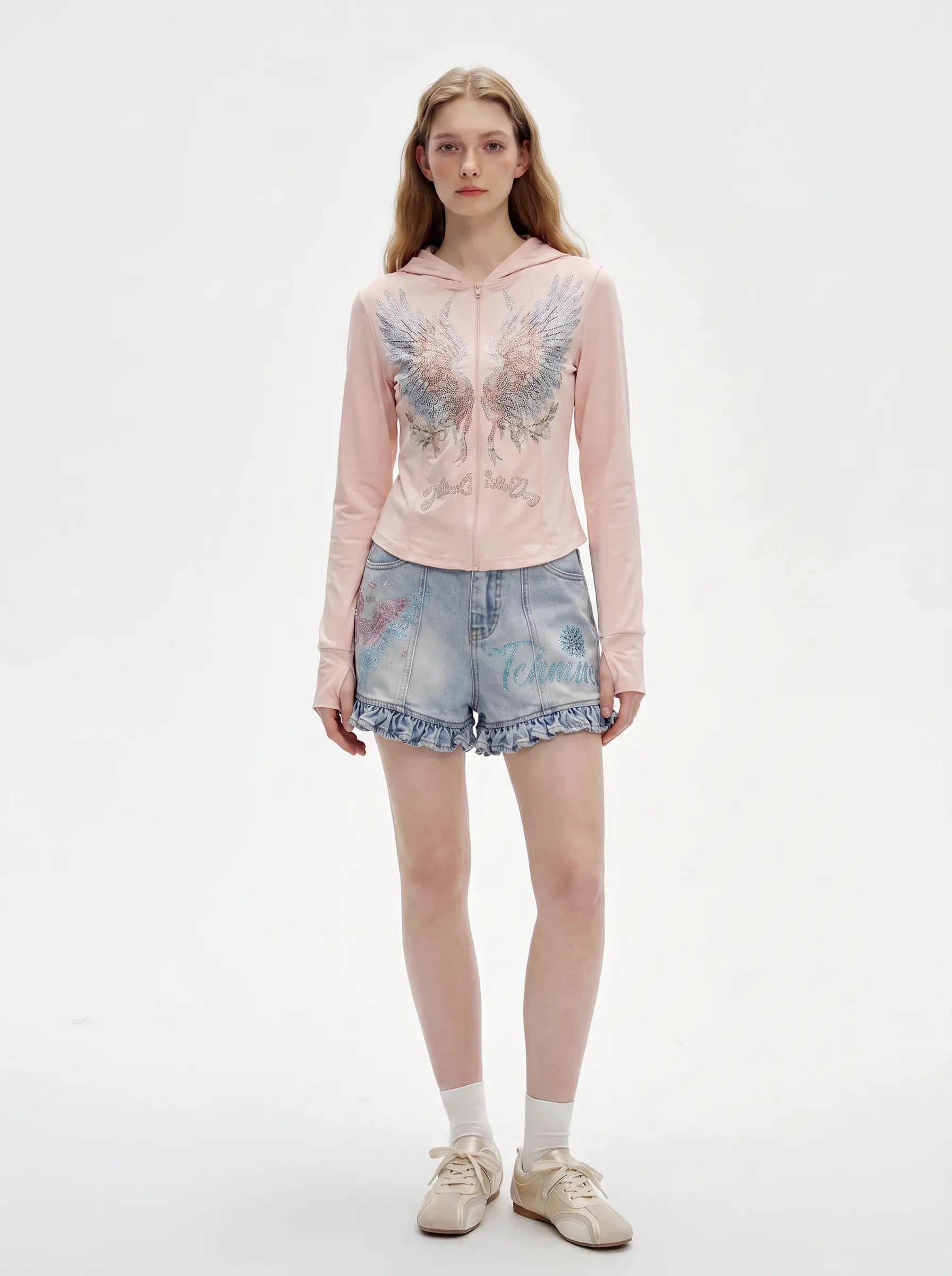 A model stands against a plain white studio background wearing a light pink long-sleeve hoodie with a large embroidered angel wing design on the front, paired with light-wash denim shorts, white socks, and beige sneakers. TCH Angel Wings Rhinestone Zip-Up Hoodie