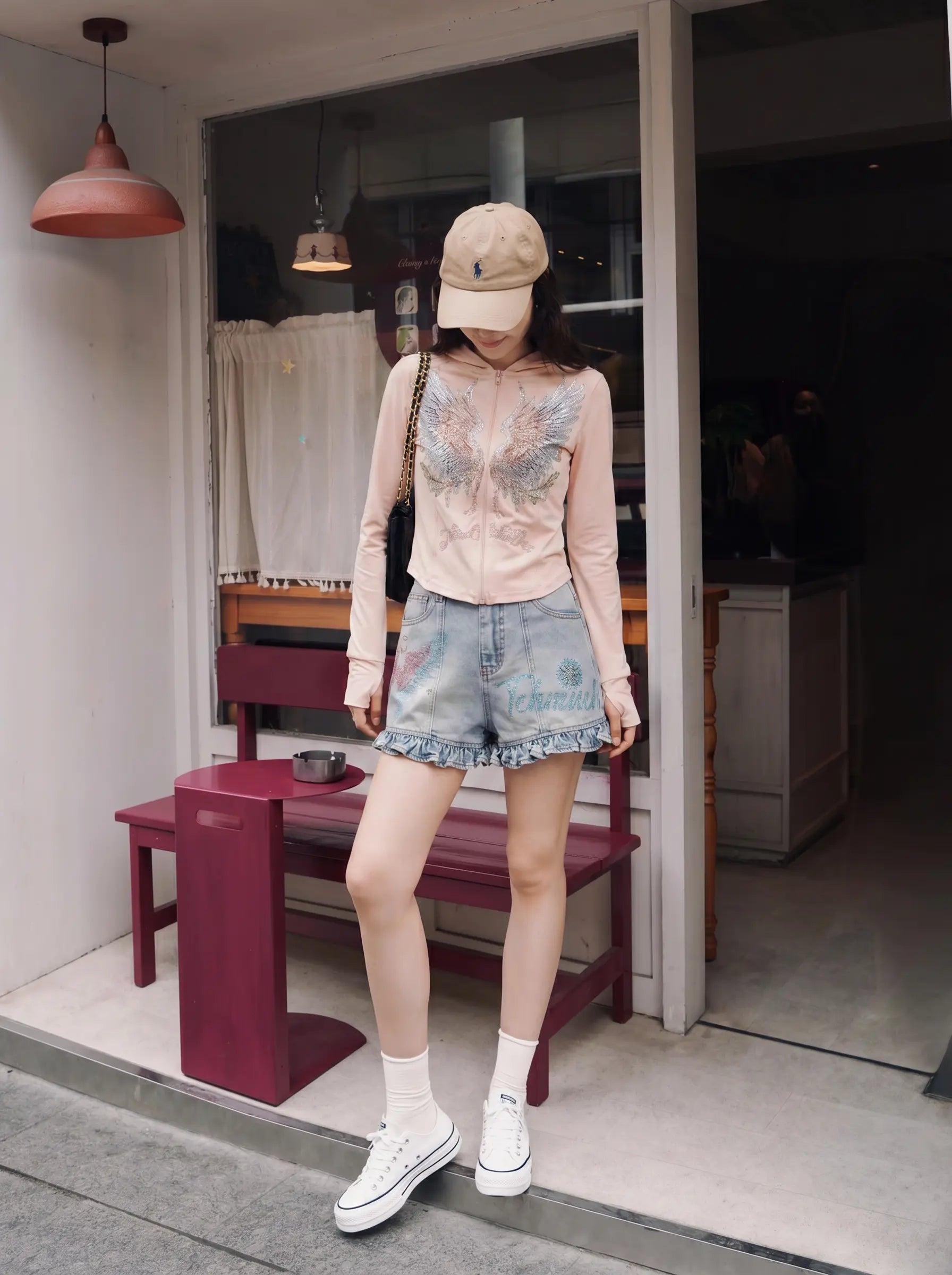 A woman stands on the threshold of a shop wearing a light pink long-sleeve crop top with a blue floral print, light-wash denim ruffle-hem shorts, white socks, and white sneakers. She is also wearing a beige baseball cap. TCH Angel Wings Rhinestone Zip-Up Hoodie