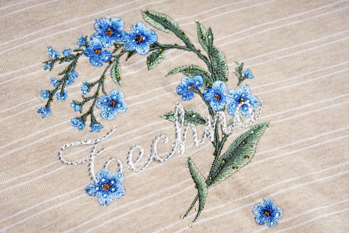 Close-up of embroidered blue forget-me-not flowers and green leaves on a beige striped fabric, with white cursive text reading 'techno' stitched across the center. TCH Beige Floral Embroidered Long Sleeve Polo Shirt
