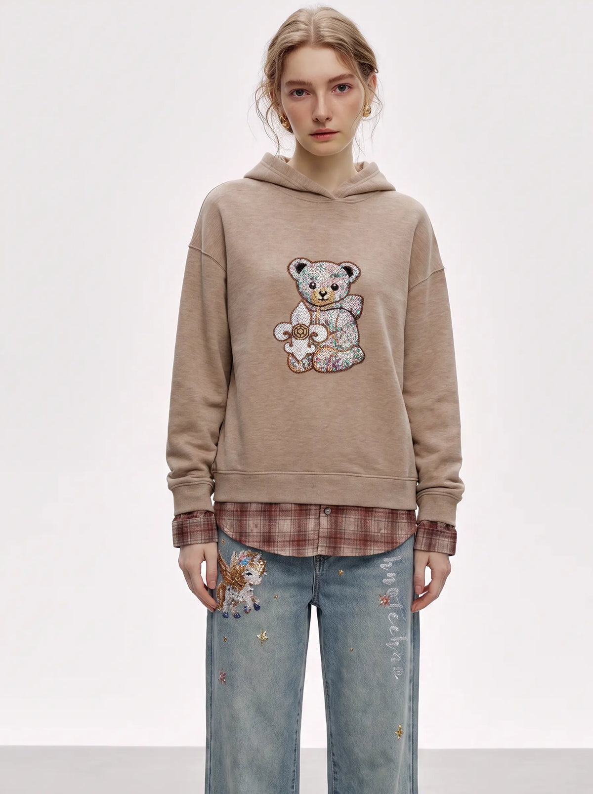 A model wearing a beige oversized hoodie with a teddy bear graphic, layered over a brown plaid shirt and paired with light blue jeans featuring a cat patch, against a plain white studio background. TCH Beige Teddy Bear Rhinestone Layered Hoodie Sweatshirt