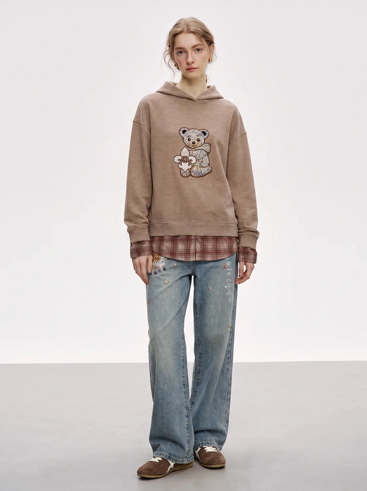 A model stands against a light gray studio background wearing a beige oversized hoodie with a teddy bear applique, layered over a brown plaid shirt, paired with light blue jeans and brown shoes. TCH Beige Teddy Bear Rhinestone Layered Hoodie Sweatshirt