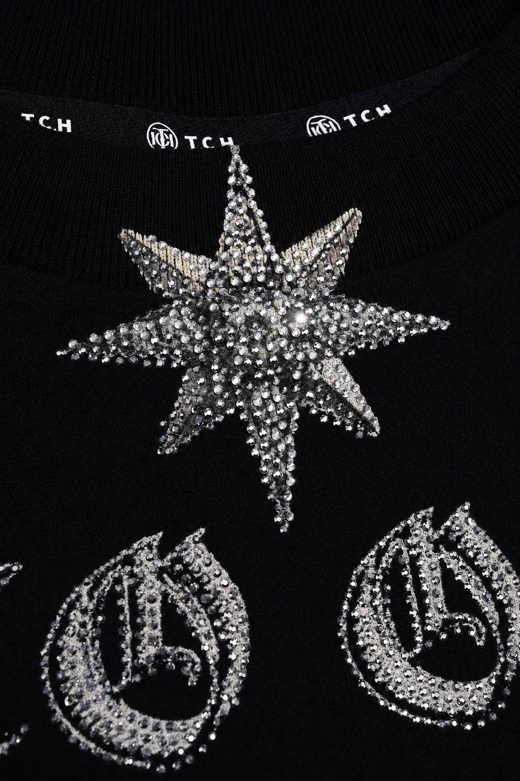 Close-up of a black knit garment featuring a large silver rhinestone star embellishment and matching silver monogram embroidery, with TCH branding visible along the inner neckband. TCH Black Old English Techno Graphic Sweatshirt