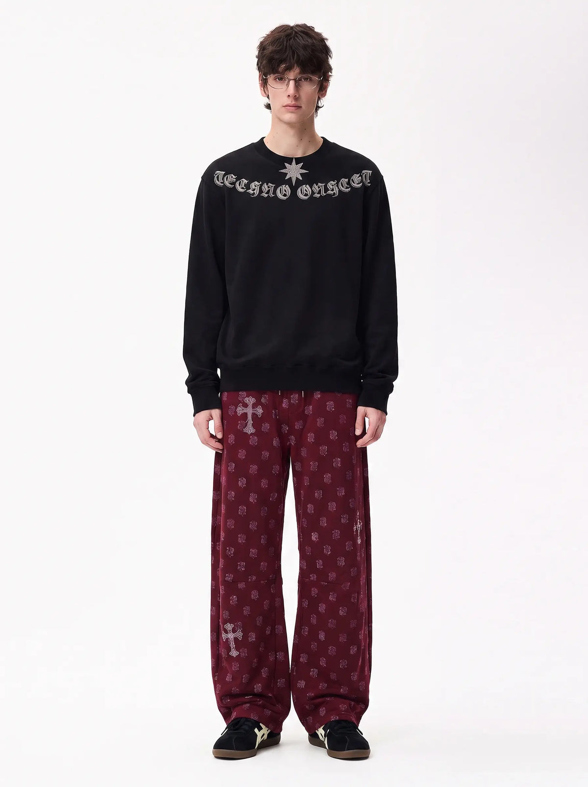 A model stands against a plain white studio background wearing a black crewneck sweatshirt with 'Technoonger' text and a star logo on the chest, paired with burgundy patterned pants and black sneakers. TCH Black Old English Techno Graphic Sweatshirt