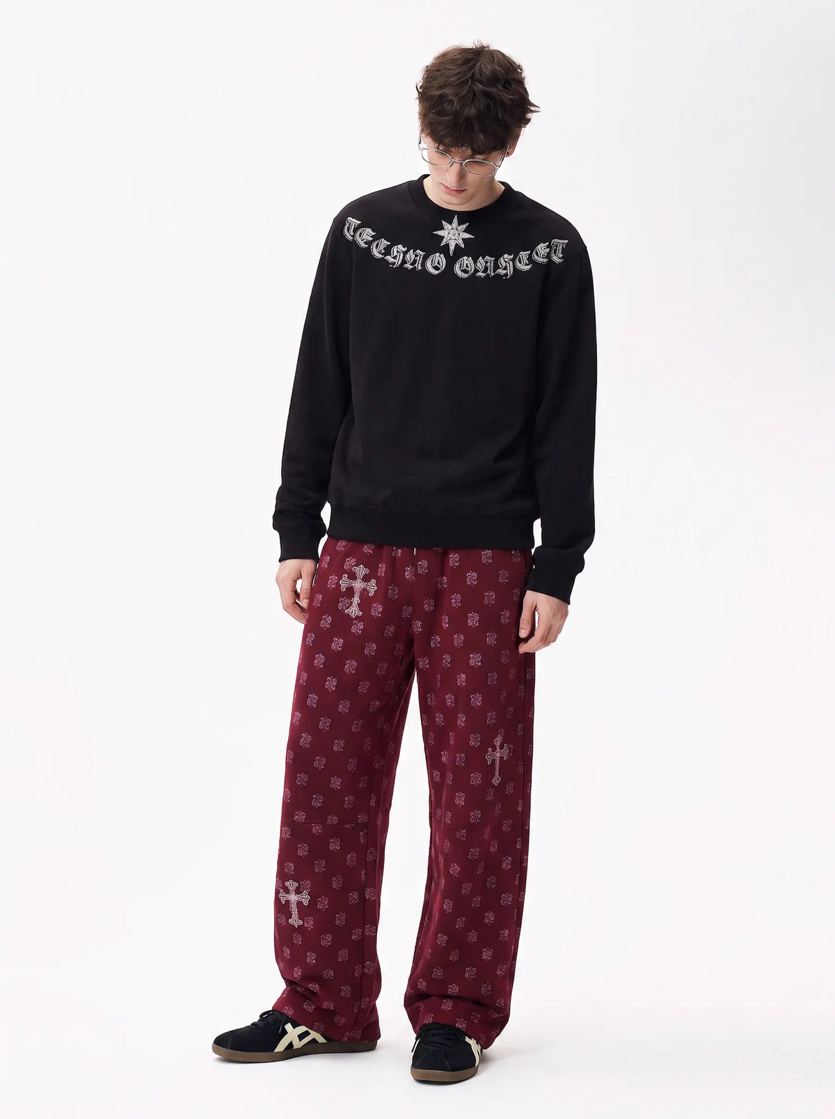 A model wearing a black crewneck sweatshirt with 'Teeth of Love' in white gothic lettering, paired with maroon patterned lounge pants and black sneakers, shot against a plain white studio background. TCH Black Old English Techno Graphic Sweatshirt