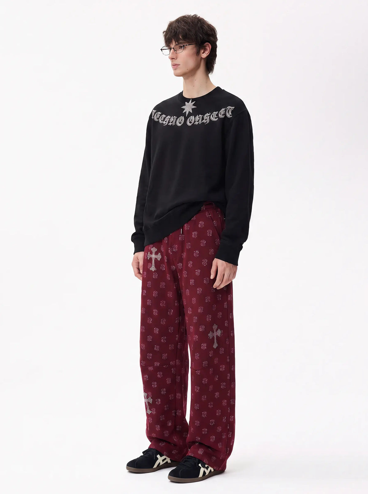 A model wearing a black crewneck sweatshirt with 'Ravage Onset' in gothic lettering and a star graphic, paired with burgundy patterned trousers and black sneakers, against a plain white studio background. TCH Black Old English Techno Graphic Sweatshirt