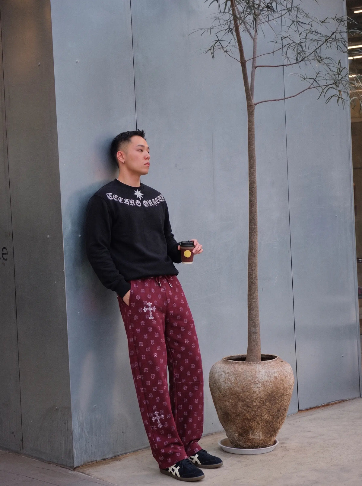 A man stands against a gray concrete wall wearing a black long-sleeve sweatshirt with white gothic lettering and maroon patterned sweatpants, holding a coffee cup next to a potted tree. TCH Black Old English Techno Graphic Sweatshirt
