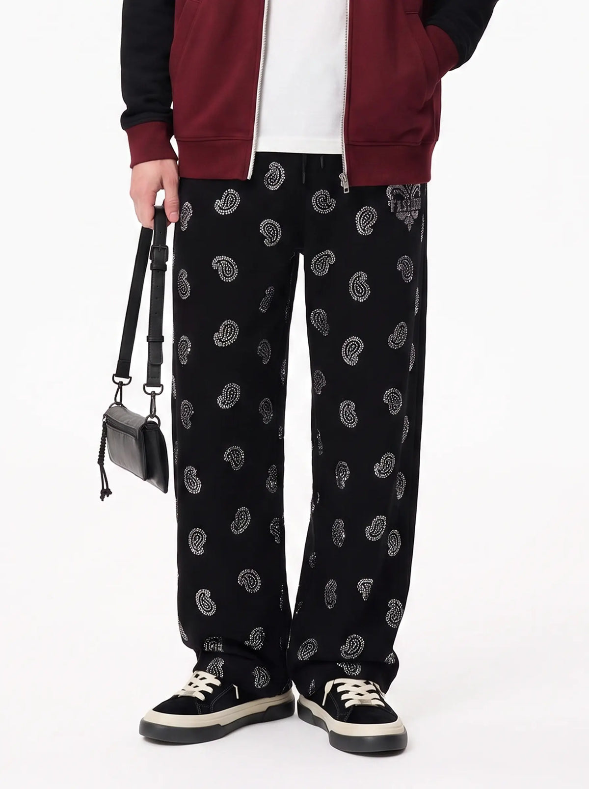 A person wearing black wide-leg pants with a repeating white paisley pattern, styled with a burgundy and black jacket, white shirt, black sneakers, and a small black shoulder bag against a plain white studio background. TCH Black Paisley Print Wide-Leg Sweatpants