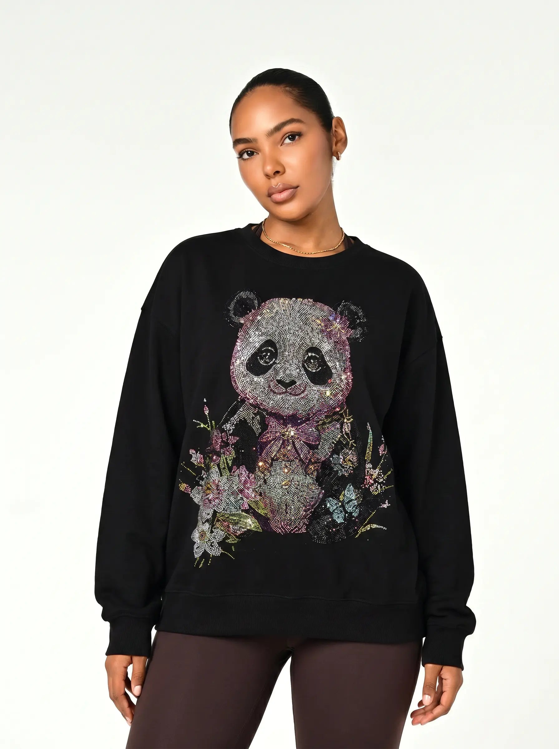 A woman modeling a black crewneck sweatshirt featuring a large, detailed graphic of a panda decorated with purple flowers and a bow, paired with dark brown leggings against a plain white studio background. TCH Black Panda Rhinestone Sweatshirt