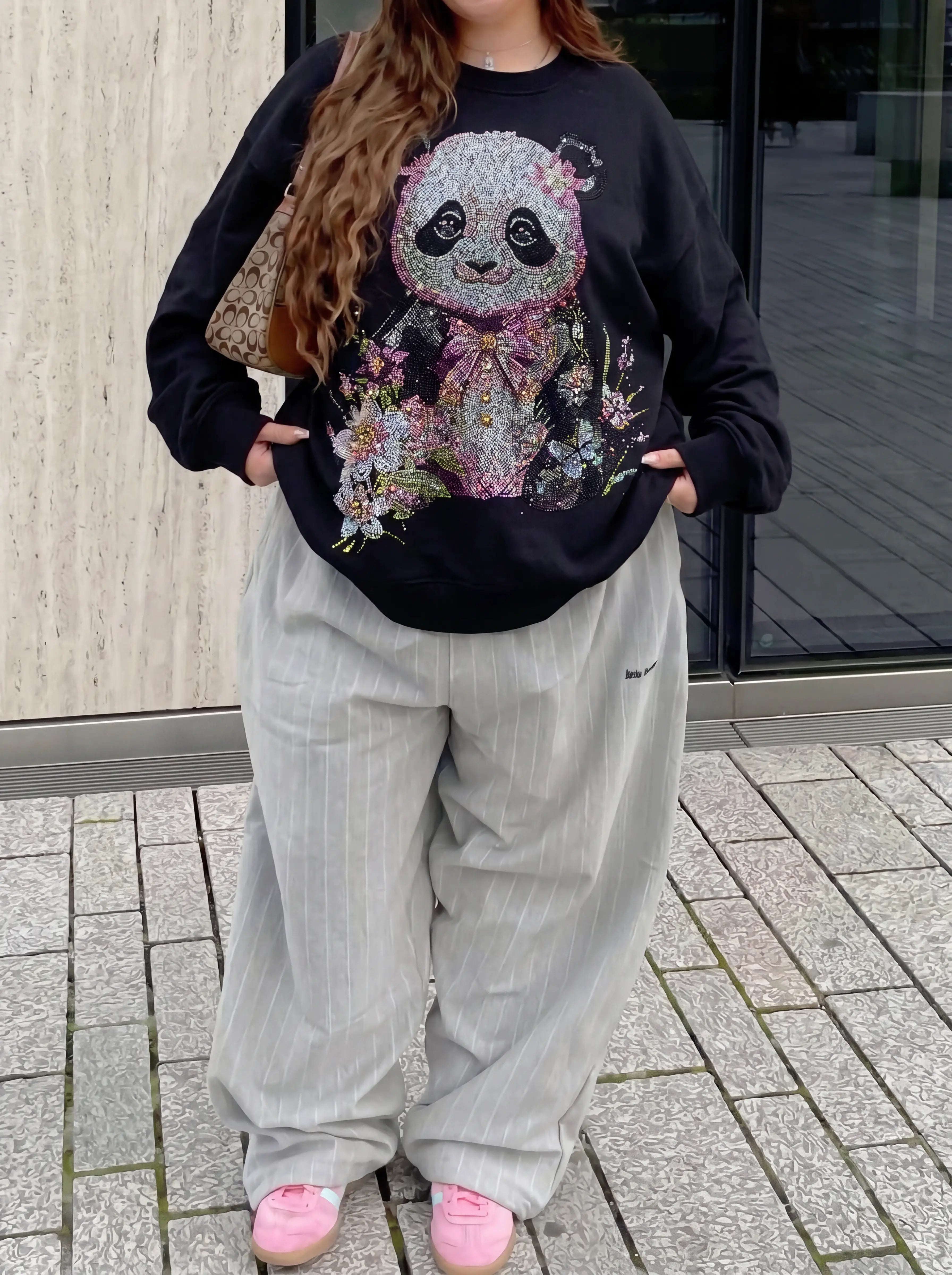 A woman wearing a black crewneck sweatshirt with a large, colorful, rhinestone-embellished panda graphic, paired with light gray pinstriped pants and pink sneakers, standing outdoors. TCH Black Panda Rhinestone Sweatshirt