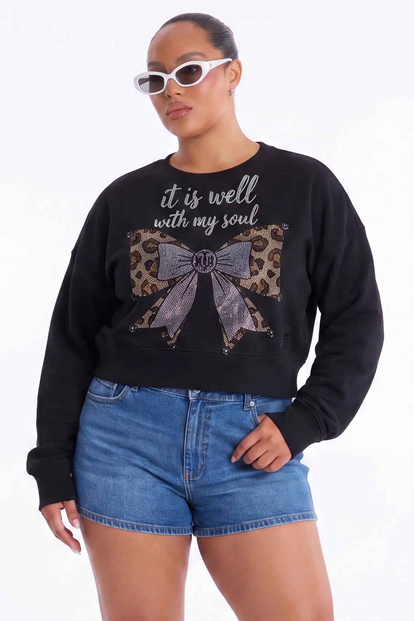 Person wearing a black sweatshirt with a bow design and text, paired with blue shorts on a white background, TCH Black Rhinestone Bow Cropped Sweatshirt