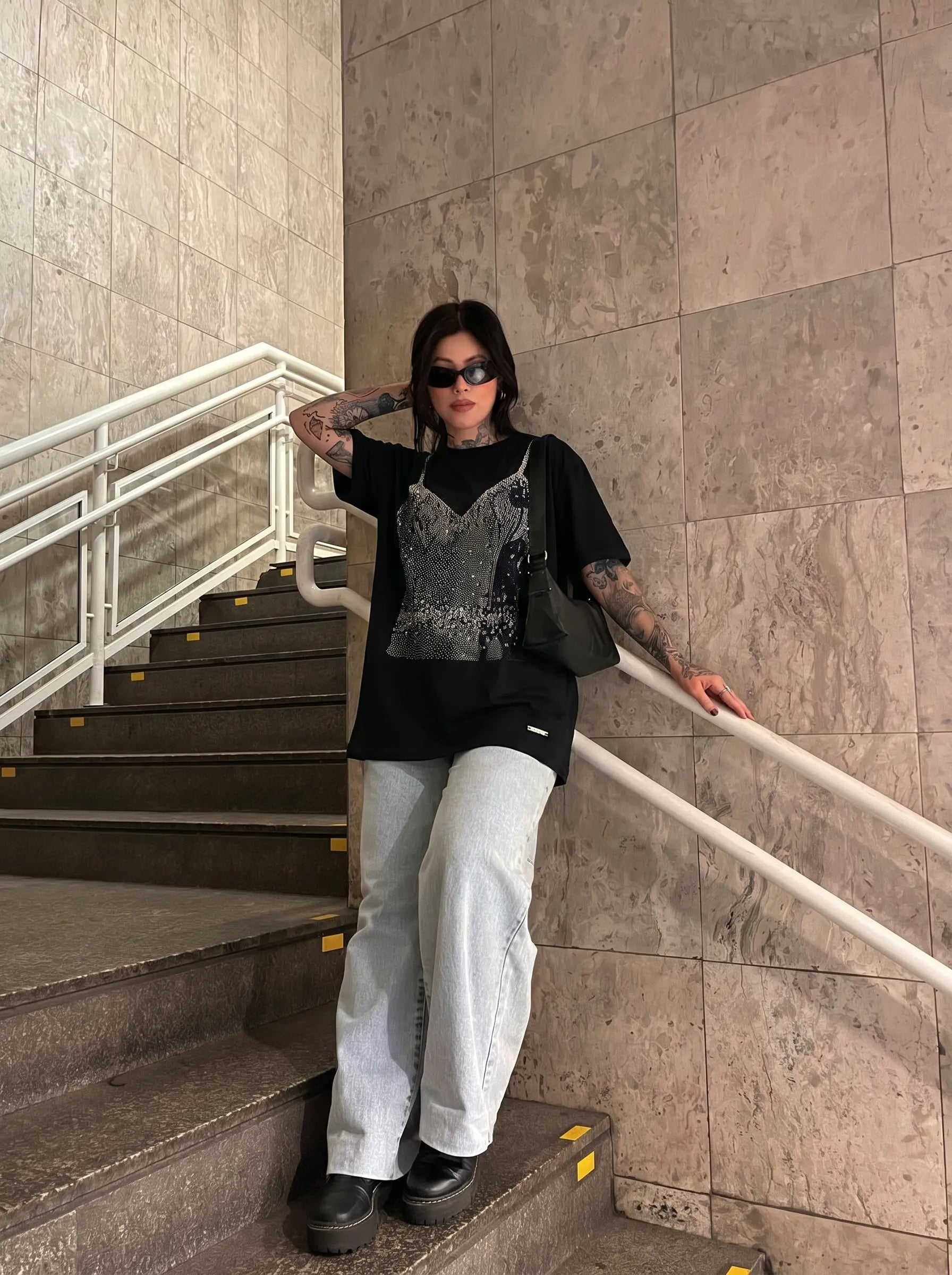 A person stands on an indoor staircase wearing a black oversized T-shirt with a silver sequin camisole layered underneath, light gray wide-leg jeans, black shoes, and black sunglasses. TCH Black Rhinestone Corset Graphic T-Shirt