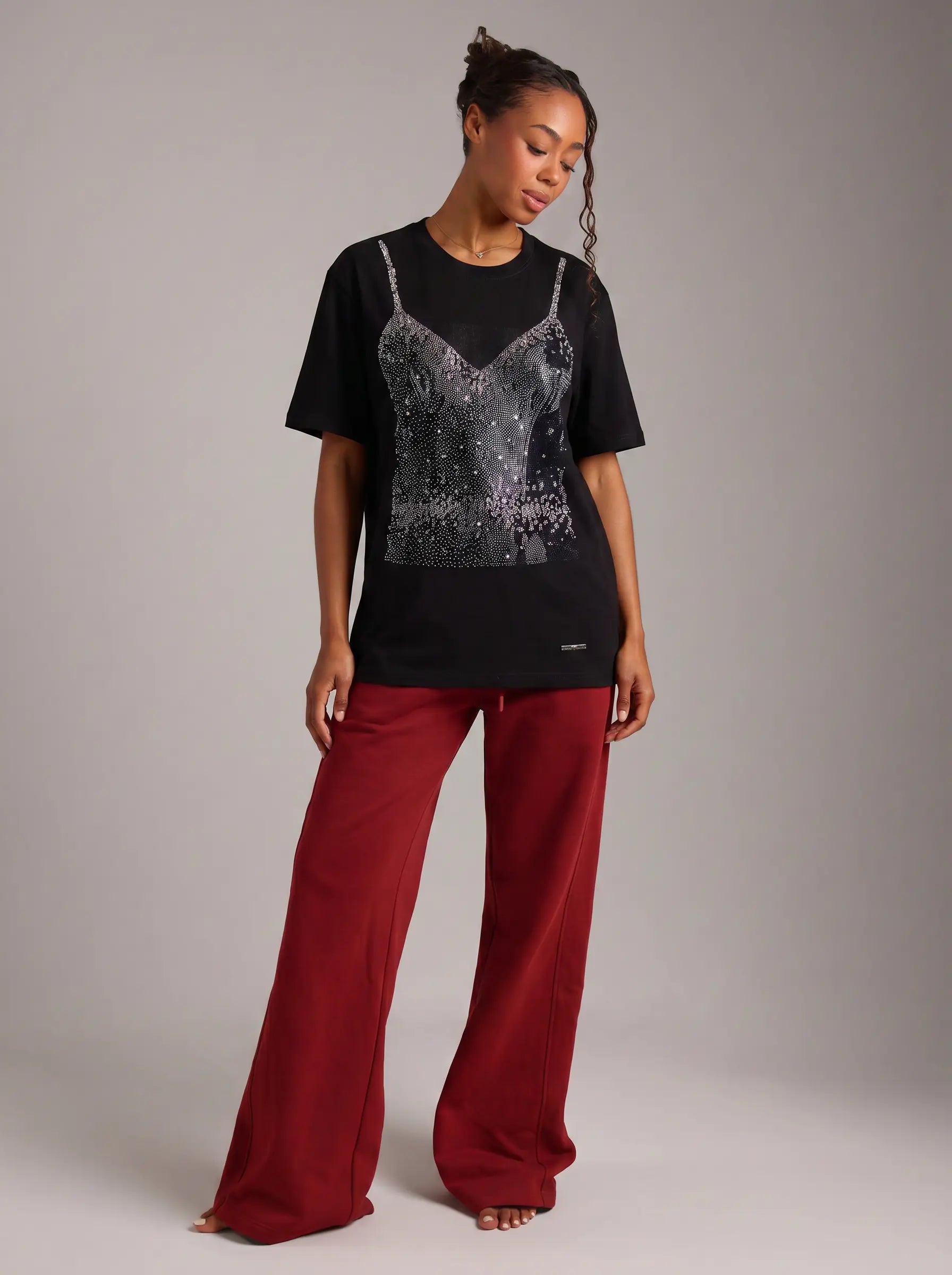 A woman modeling a black short-sleeve t-shirt with a silver sequined spaghetti-strap bodice overlay, paired with wide-leg burgundy pants, against a neutral gray studio background. TCH Black Rhinestone Corset Graphic T-Shirt