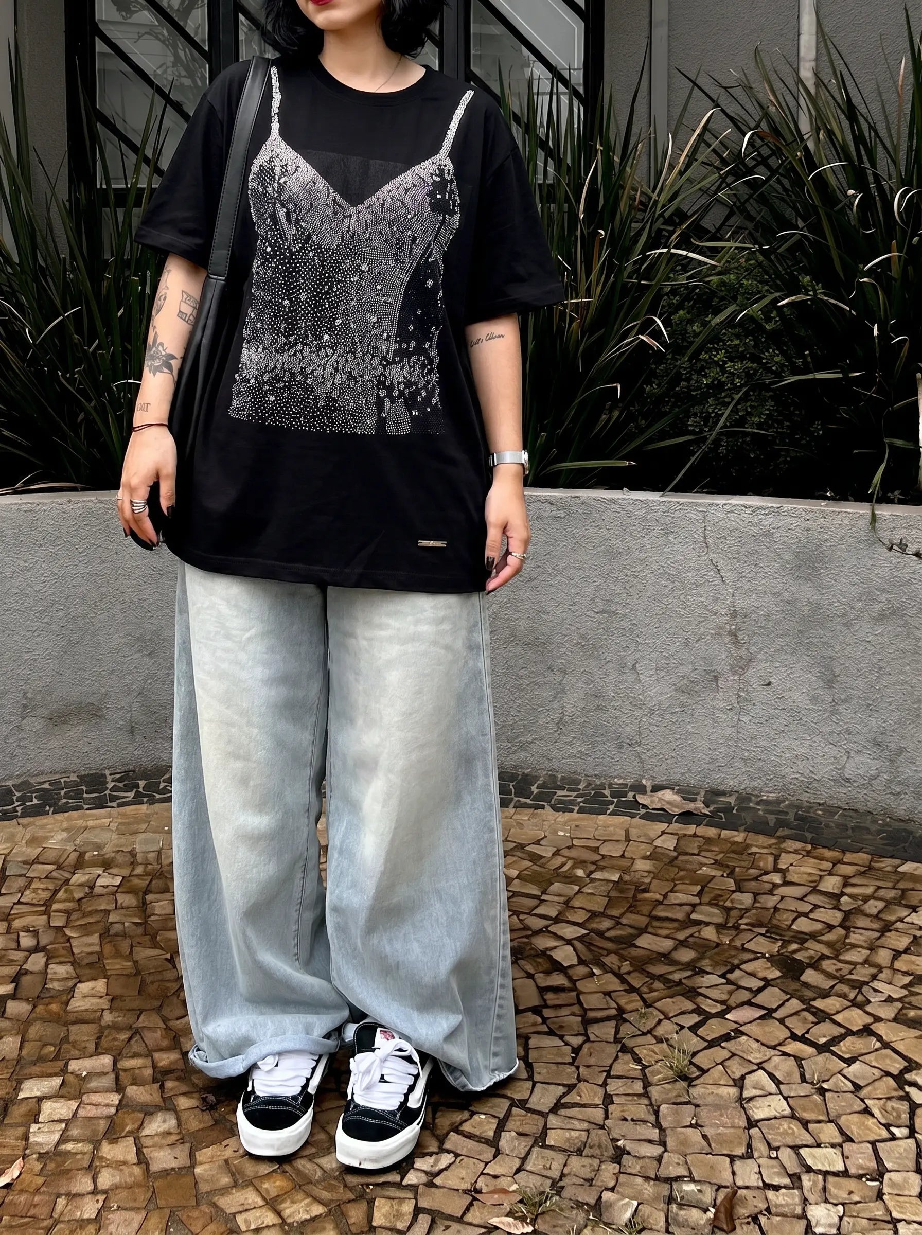 A person wearing a black graphic t-shirt with a silver rhinestone-embellished camisole print, paired with wide-leg light-wash jeans and black-and-white sneakers, standing outdoors. TCH Black Rhinestone Corset Graphic T-Shirt