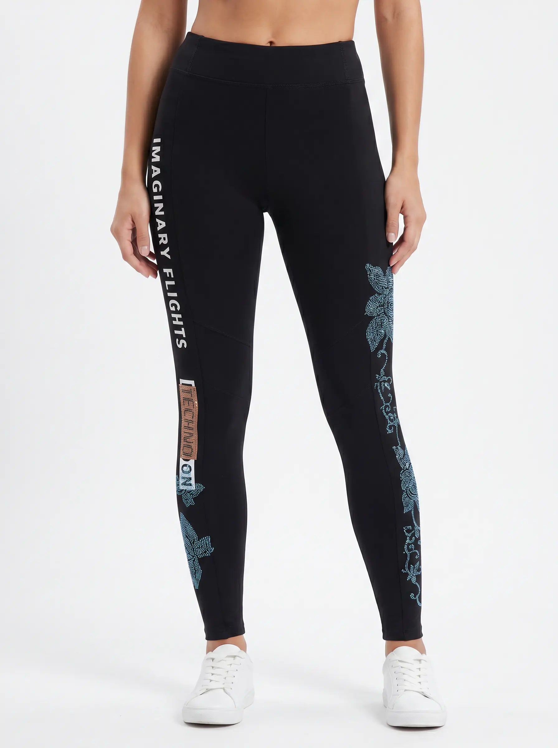A model wearing black high-waisted leggings with white vertical text reading 'IMAGINARY FLIGHTS' and blue floral graphics down the sides, paired with white sneakers against a plain white studio background. TCH Black Rhinestone Floral Leggings
