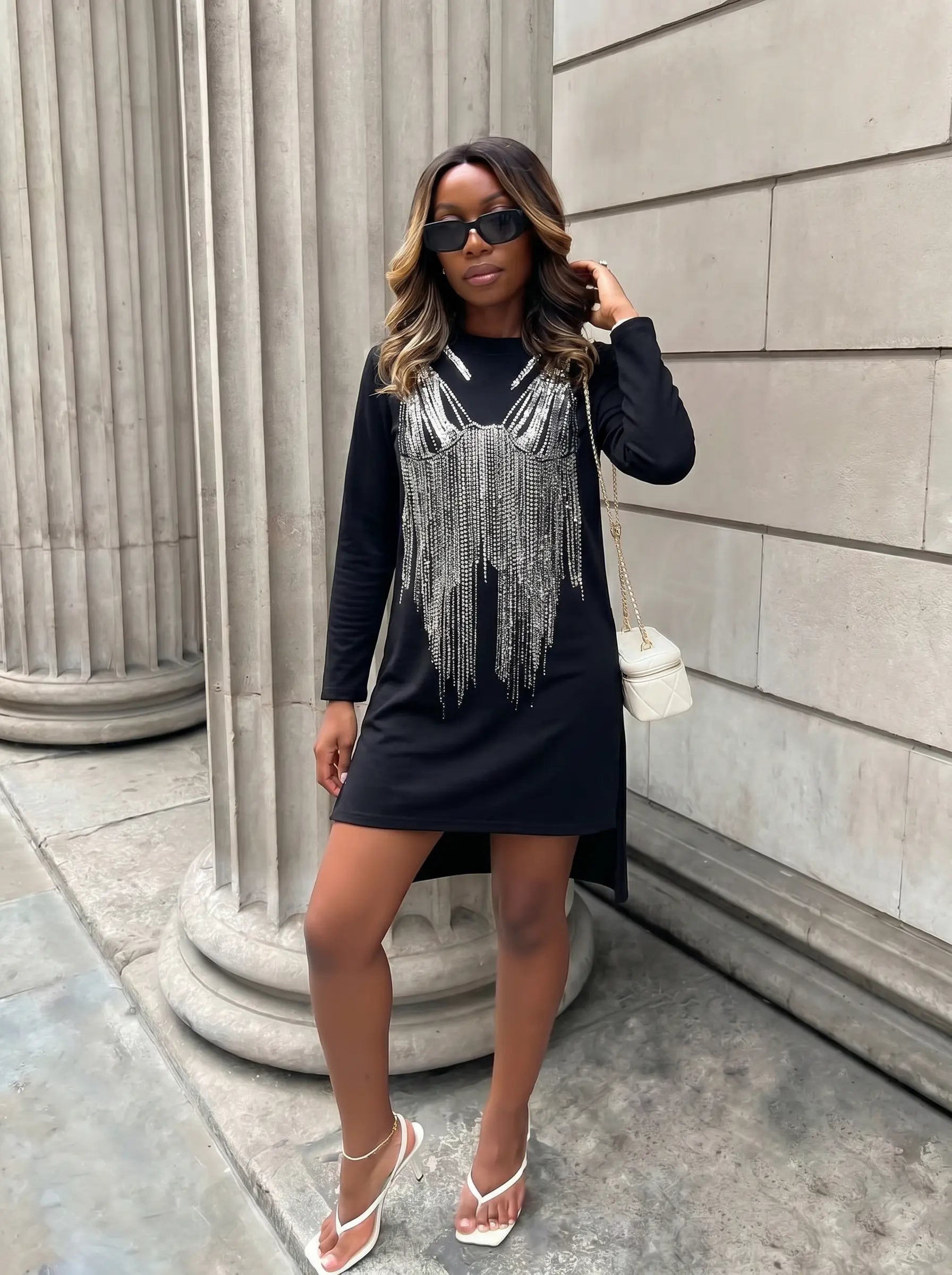A woman models a black long-sleeve mini dress with a silver fringe embellishment across the chest, styled with a small white shoulder bag and white sandals, standing outdoors next to a stone column. TCH Black Rhinestone Fringe Detail Long Sleeve Mini Dress