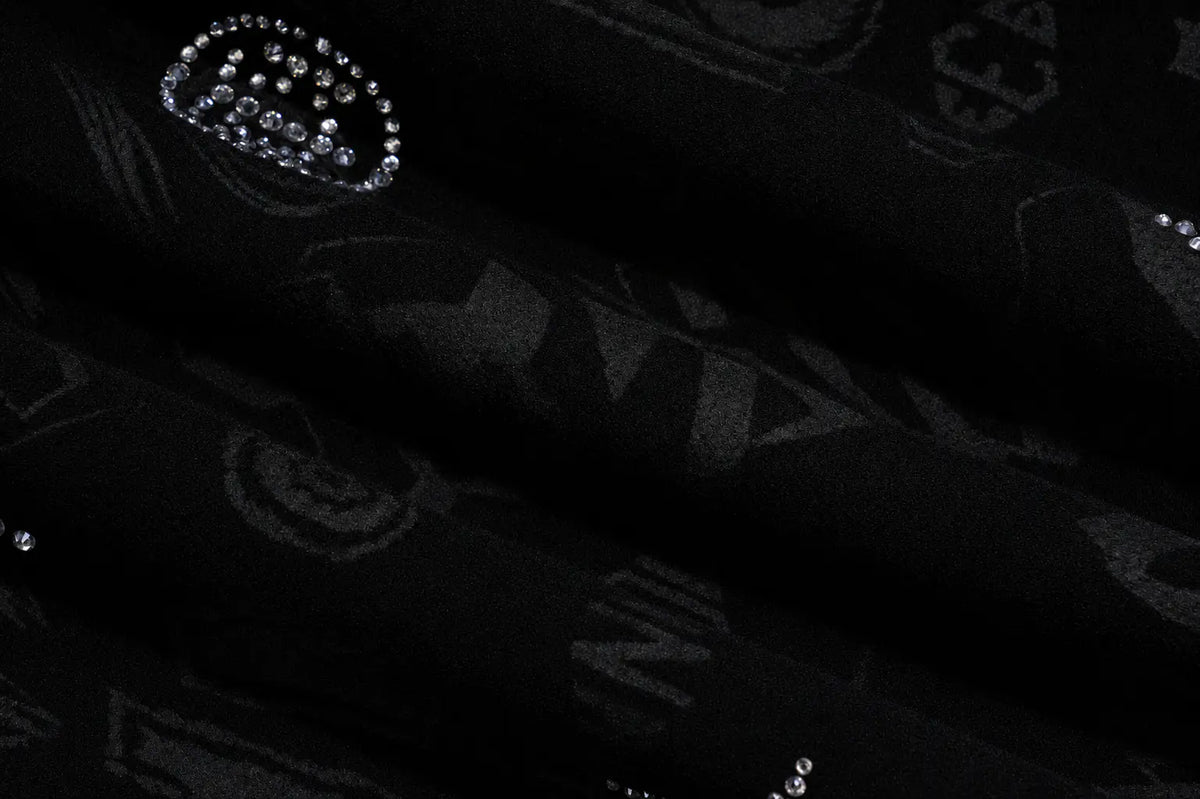 A close-up of a black fabric with a subtle tonal pattern and a decorative silver rhinestone crown embellishment. TCH Black Rhinestone Monogram Zip Up Hoodie Jacket