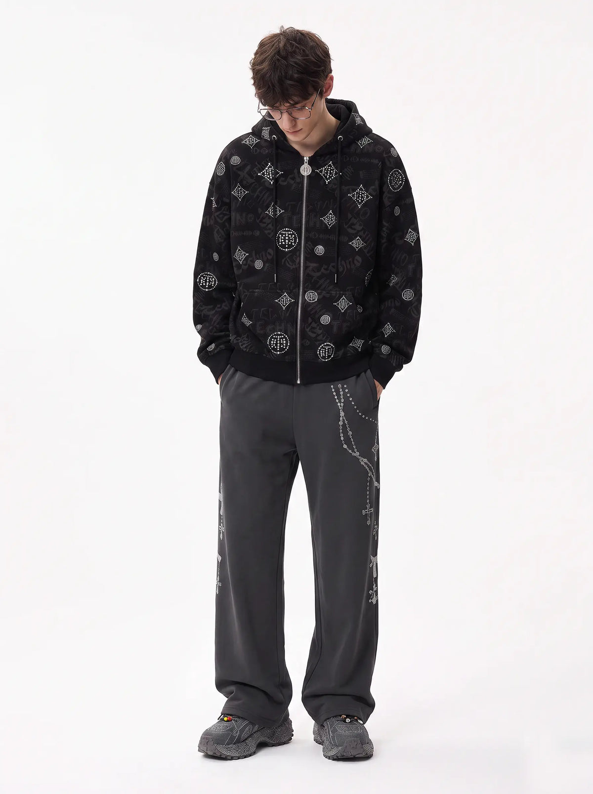 A man wearing a black zip-up hoodie with a silver-tone chain and geometric pattern, paired with dark gray sweatpants and gray sneakers, standing against a plain white studio background. TCH Black Rhinestone Monogram Zip Up Hoodie Jacket