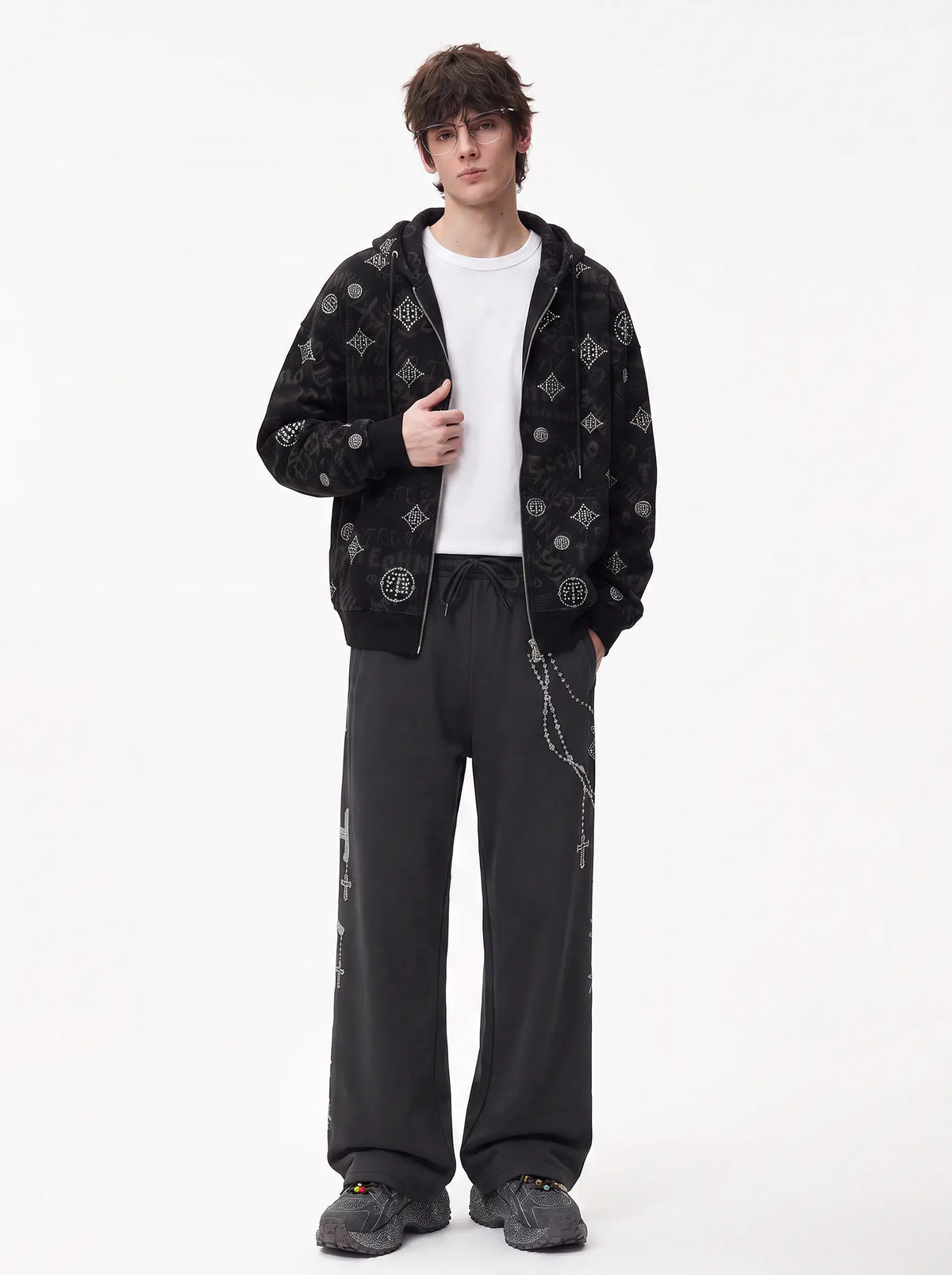 A model stands against a plain white studio background wearing a black zip-up hoodie with white graphic patterns, a white t-shirt, loose black sweatpants, and gray sneakers. TCH Black Rhinestone Monogram Zip Up Hoodie Jacket