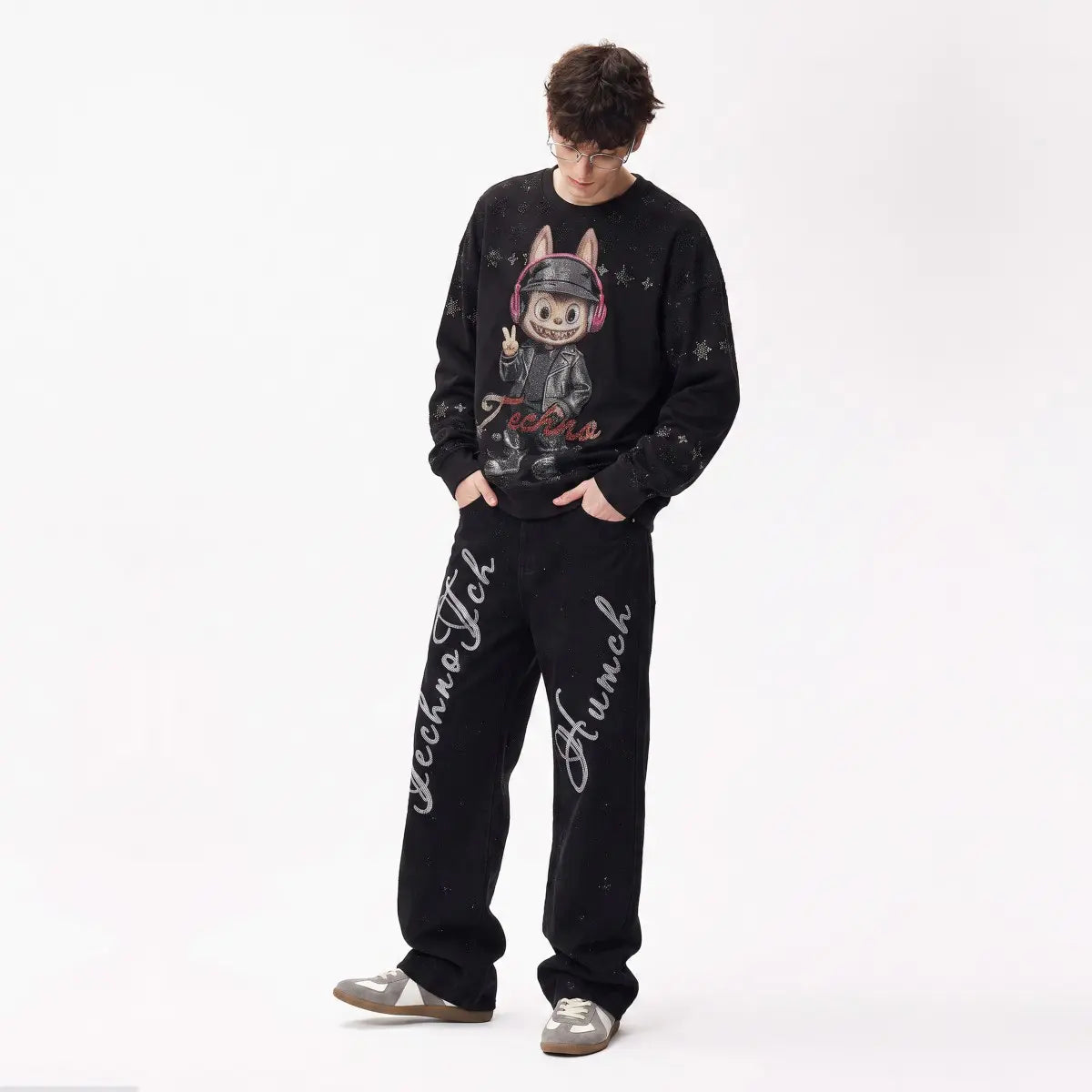 A model wearing a black crewneck sweatshirt and matching sweatpants set. The sweatshirt features a cartoon rabbit graphic wearing headphones with 'Techno Hunch' written in red. TCH Black Techno Bunny Rhinestone Graphic Sweatshirt