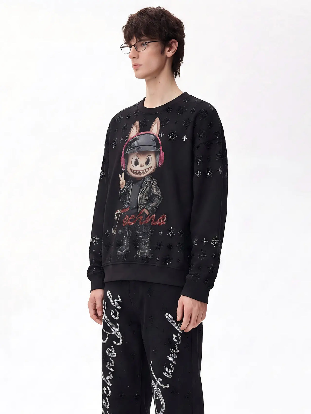 A model wearing a black Techno Fich branded crewneck sweatshirt with a cartoon graphic and matching black sweatpants with white script lettering, set against a plain white studio background. TCH Black Techno Bunny Rhinestone Graphic Sweatshirt
