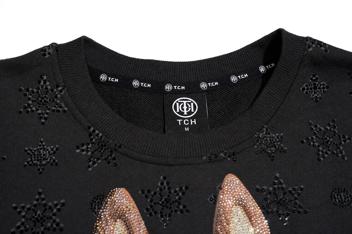 Close-up of a black crewneck sweater with rhinestone snowflake patterns and rhinestone-embellished bunny ears at the bottom, featuring a visible TCH neck label. TCH Black Techno Bunny Rhinestone Graphic Sweatshirt