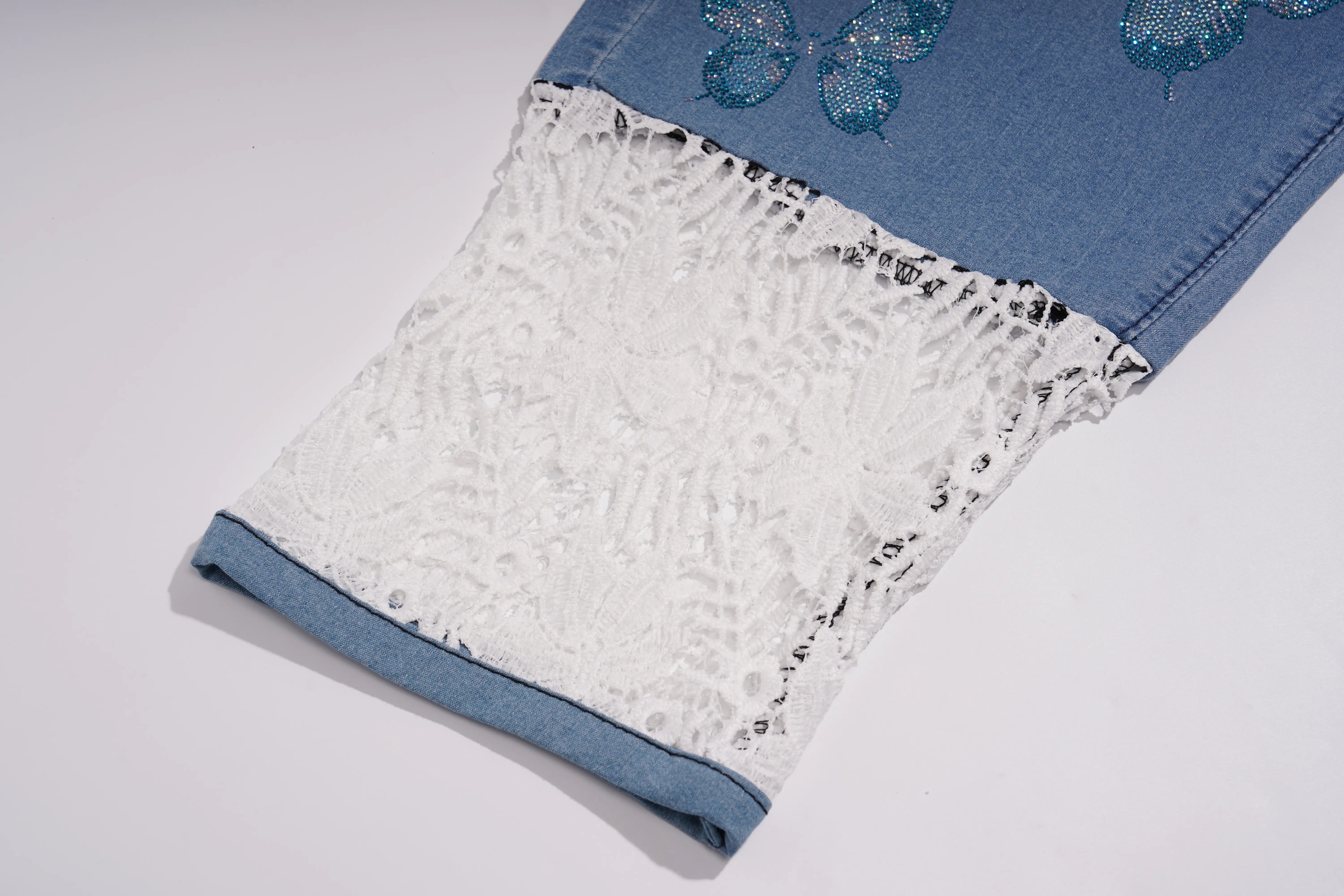 Close-up of a denim garment, likely a skirt or pants leg, featuring a wide white lace panel with sequined edges and blue butterfly embellishments near the top on a light gray background. TCH Blue Butterfly & Teddy Bear Lace Hem Denim Jeans