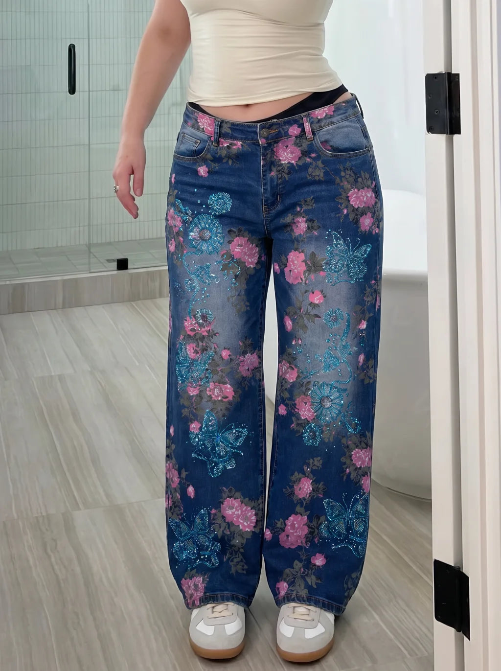 A person wearing wide-leg blue denim jeans with a pink and blue floral and butterfly print, paired with a beige crop top and white sneakers, standing in a bathroom. TCH Blue Floral Butterfly Rhinestone Wide-Leg Jeans