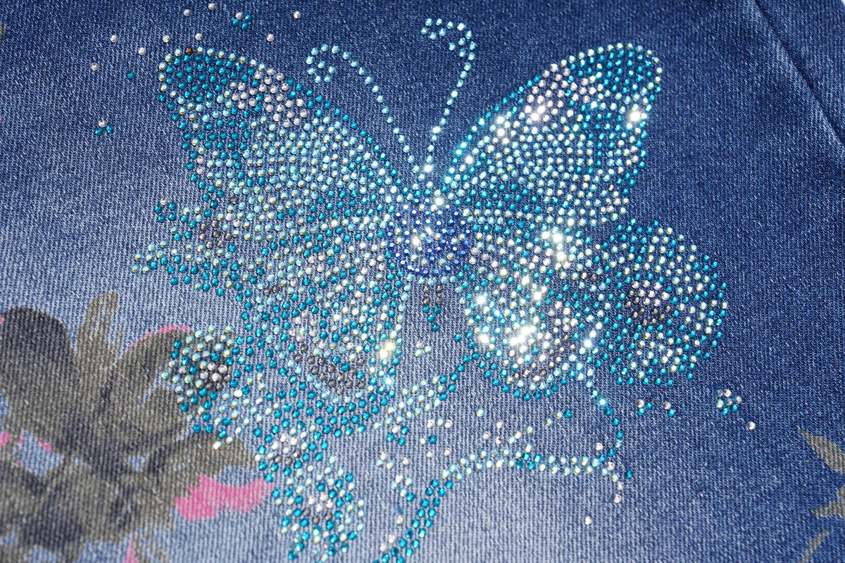 A close-up of a blue denim fabric featuring a large butterfly embellished with sparkling rhinestones in shades of blue, white, and silver. TCH Blue Floral Butterfly Rhinestone Wide-Leg Jeans