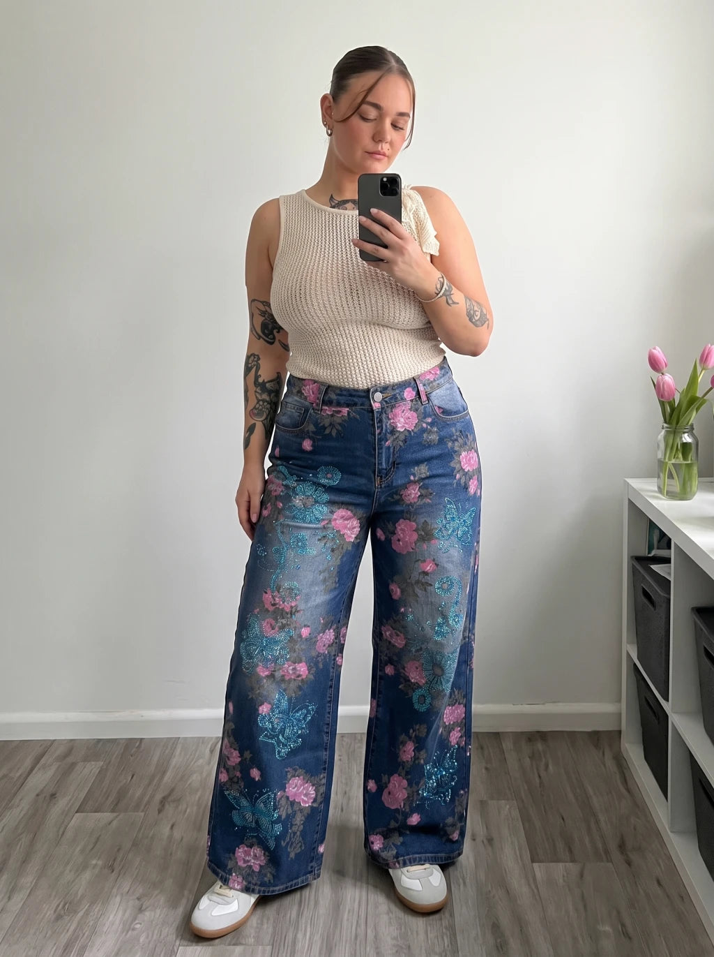 A woman takes a mirror selfie wearing high-waisted, wide-leg blue denim jeans with a pink and teal floral print, paired with a cream ribbed tank top and white sneakers. TCH Blue Floral Butterfly Rhinestone Wide-Leg Jeans