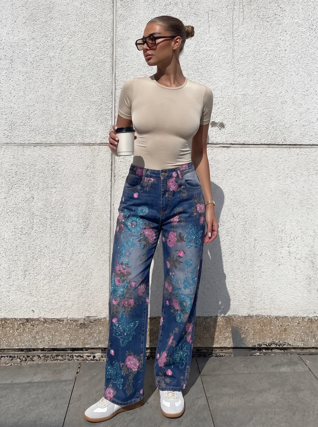 A woman stands outdoors against a light concrete wall wearing a beige short-sleeve fitted top, blue floral wide-leg jeans, white sneakers, and sunglasses, while holding a takeaway coffee cup. TCH Blue Floral Butterfly Rhinestone Wide-Leg Jeans