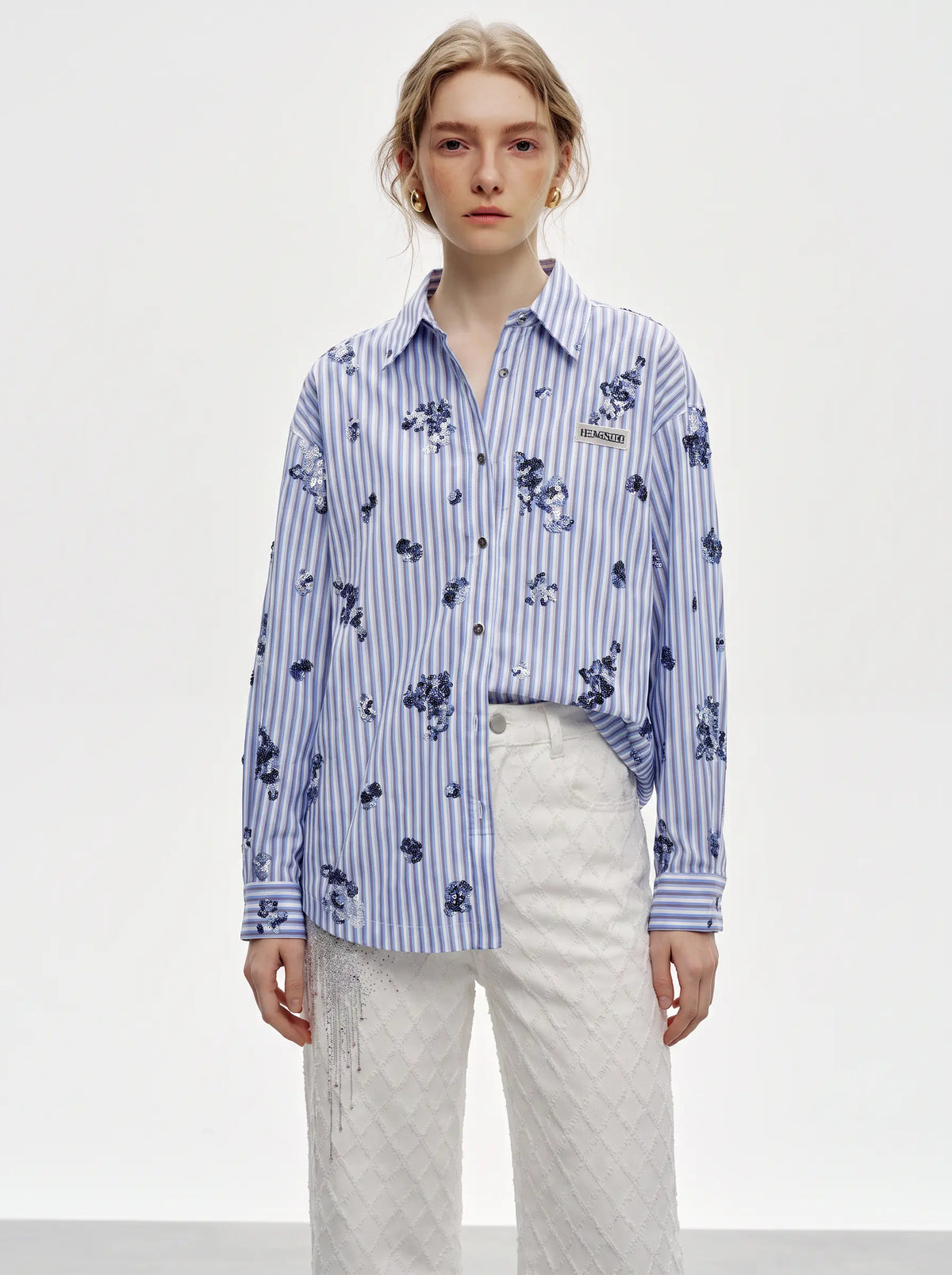 A model wearing a blue and white striped button-up shirt with a dark blue floral print, tucked into white textured pants against a plain white studio background. TCH Blue Floral Embroidered Pinstripe Shirt
