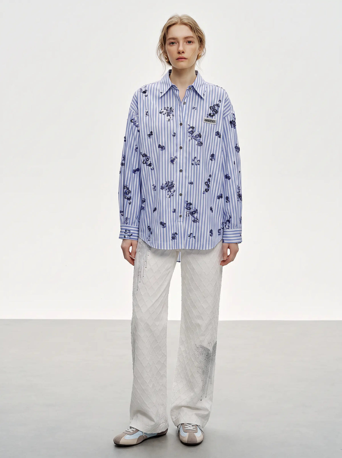 A model stands in a studio wearing a blue and white vertically striped button-up shirt with a dark blue floral print, paired with light gray wide-leg trousers and silver sneakers. TCH Blue Floral Embroidered Pinstripe Shirt