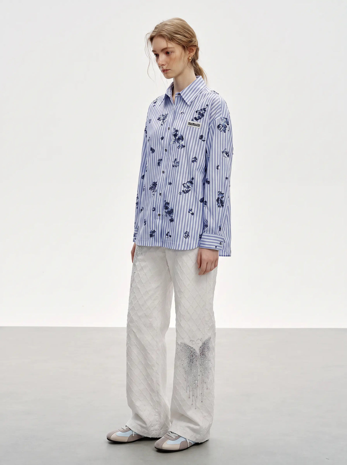 A model stands in a studio wearing a blue and white striped button-up shirt with a dark floral print, paired with wide-leg white trousers and light-colored sneakers. TCH Blue Floral Embroidered Pinstripe Shirt