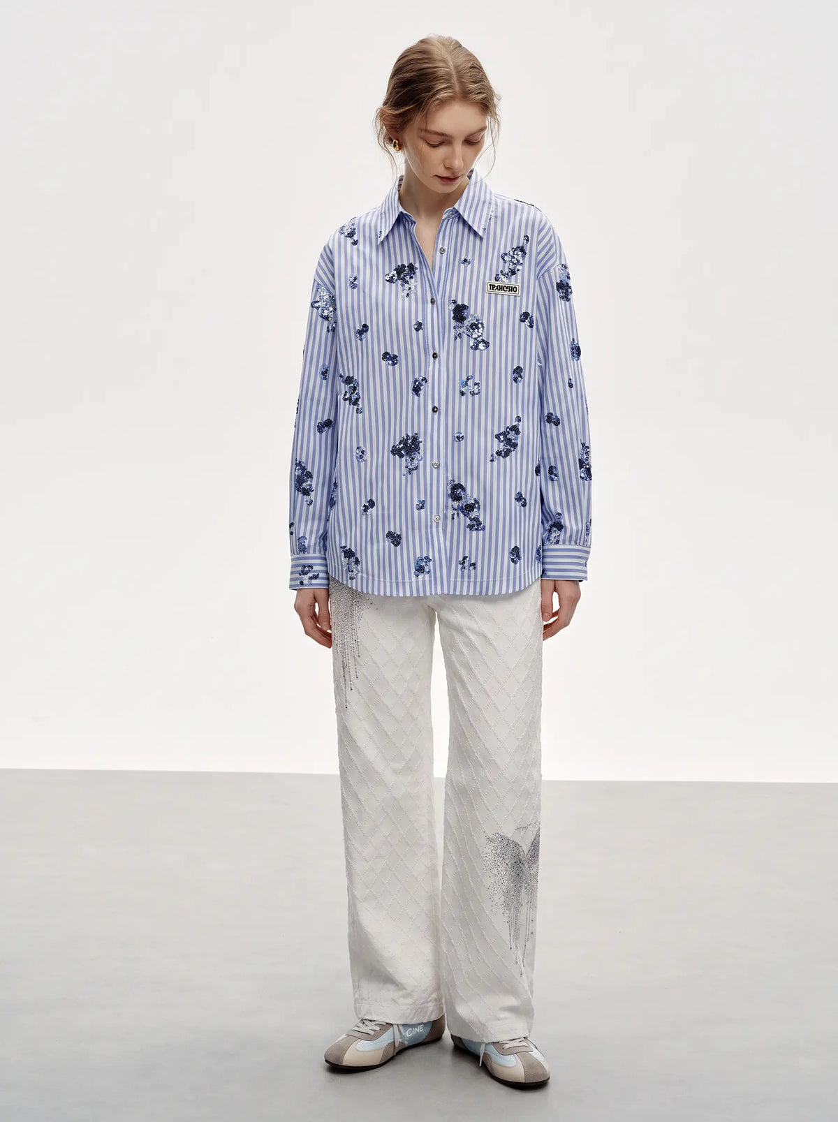 A model stands in a studio wearing a light blue and white vertically striped button-up shirt with a dark blue floral print, paired with off-white wide-leg trousers and gray sneakers. TCH Blue Floral Embroidered Pinstripe Shirt