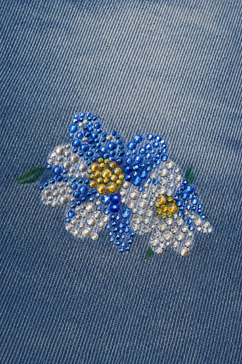 A close-up of a blue denim fabric featuring an embroidered floral patch with blue, white, and yellow rhinestone flowers and small green leaves. TCH Blue Floral Rhinestone Wide-Leg Jeans