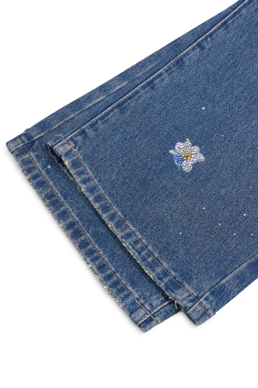 Close-up of folded blue denim jeans showing the fabric texture, stitching, and a small embroidered flower detail on a white background. TCH Blue Floral Rhinestone Wide-Leg Jeans