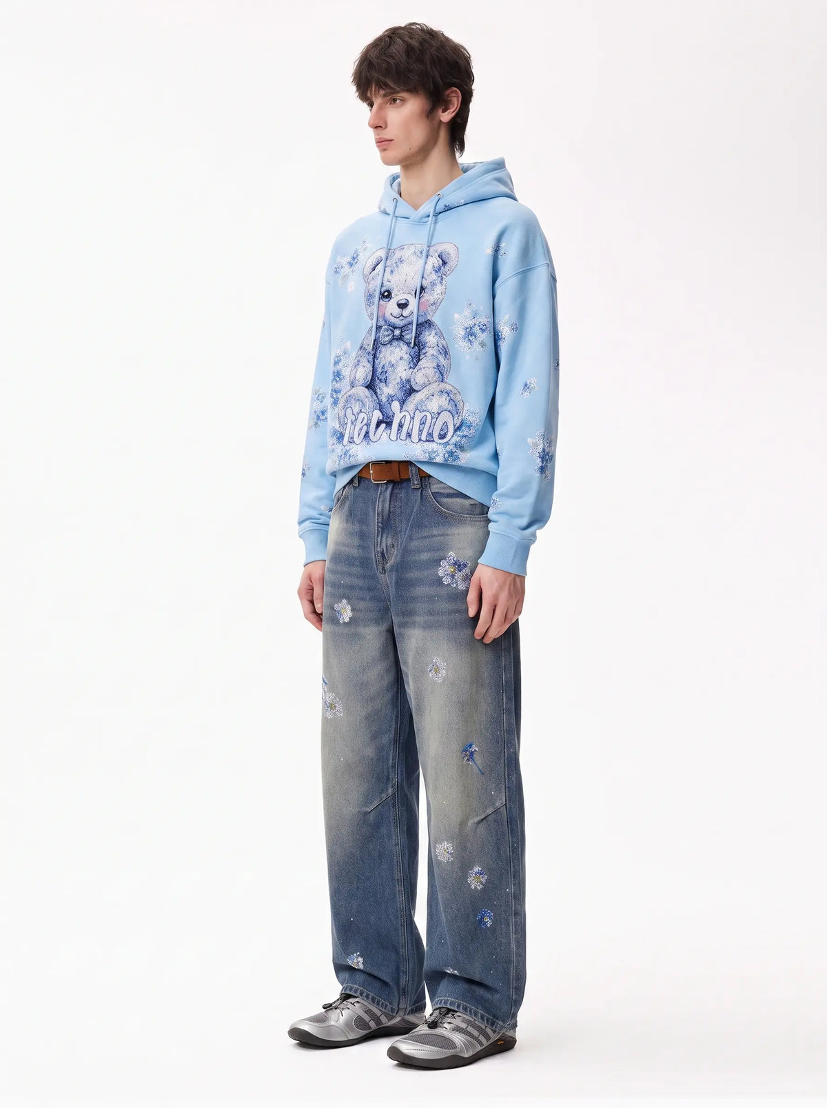 A model stands against a plain white studio background wearing a light blue hoodie with a teddy bear graphic and the word 'Felicity' on the front, paired with distressed blue jeans and gray sneakers. TCH Blue Floral Rhinestone Wide-Leg Jeans