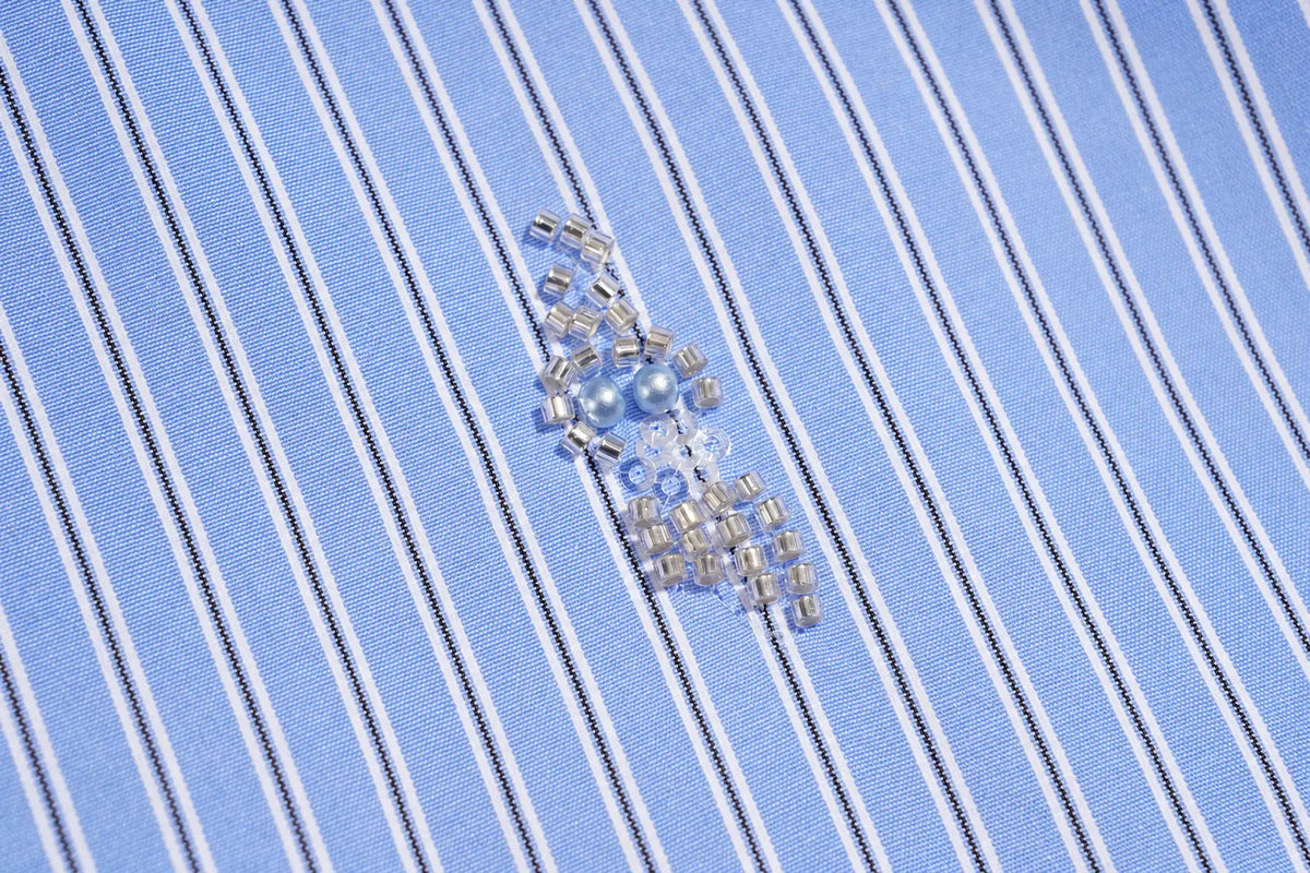 A close-up of a light blue and white striped fabric with a small pile of clear and silver seed beads placed in the center. TCH Blue Pinstripe Embroidered Pocket Shirt