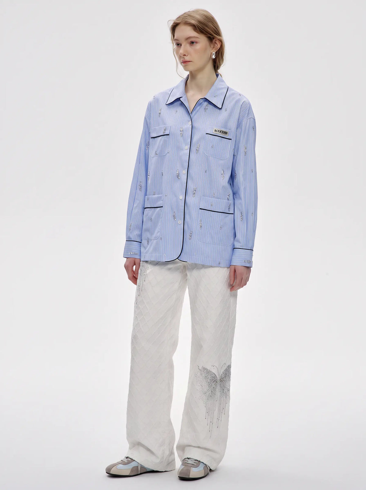 A model stands against a light gray studio background wearing a light blue long-sleeve button-up shirt with subtle vertical stripes and a small chest patch, paired with off-white wide-leg pants and gray slip-on shoes. TCH Blue Pinstripe Embroidered Pocket Shirt