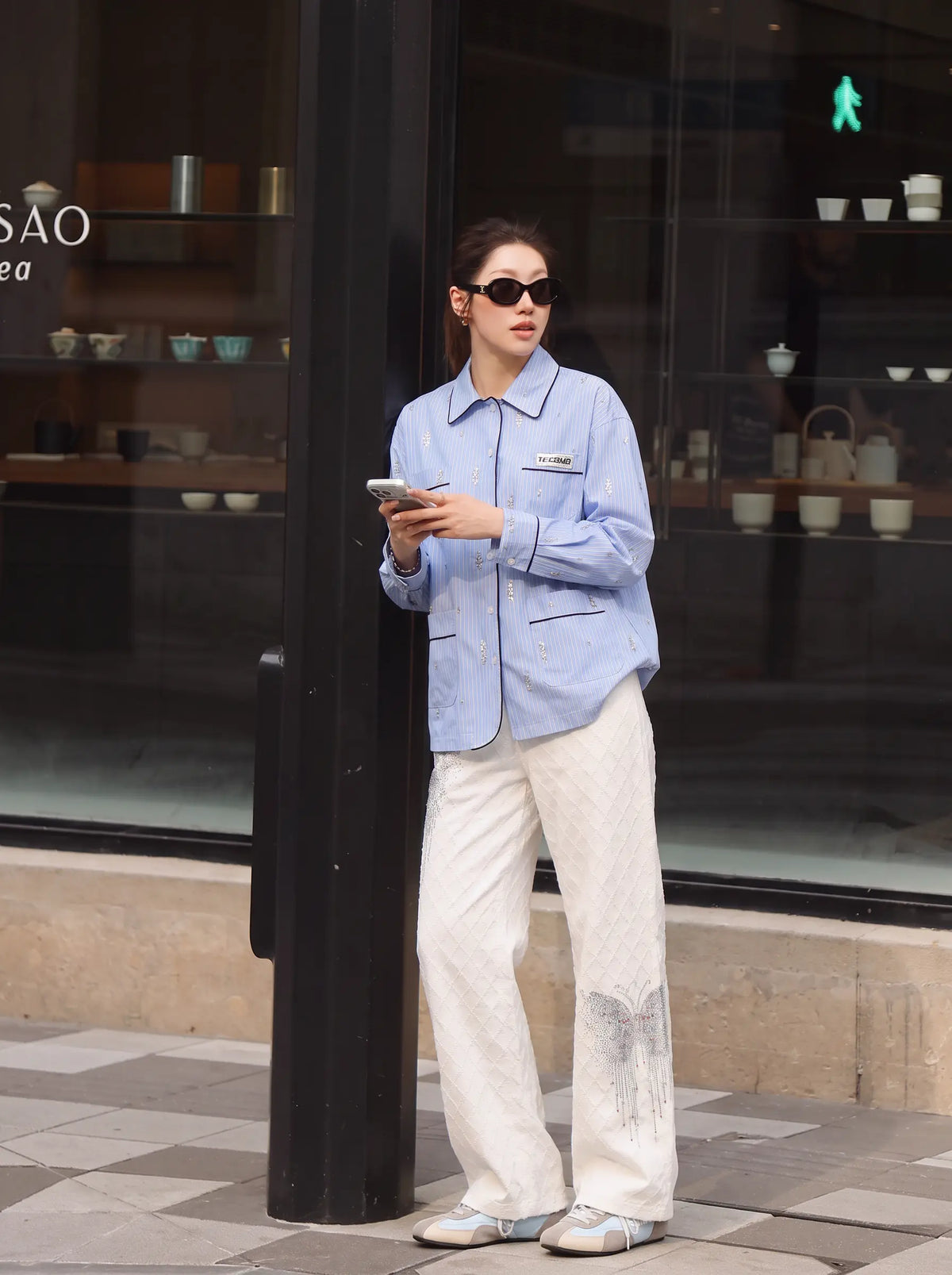 A woman stands outdoors in front of a storefront window wearing a light blue button-up shirt with dark collar and cuff trim, paired with off-white wide-leg trousers and light-colored shoes. She is holding a phone and wearing dark sunglasses. TCH Blue Pinstripe Embroidered Pocket Shirt
