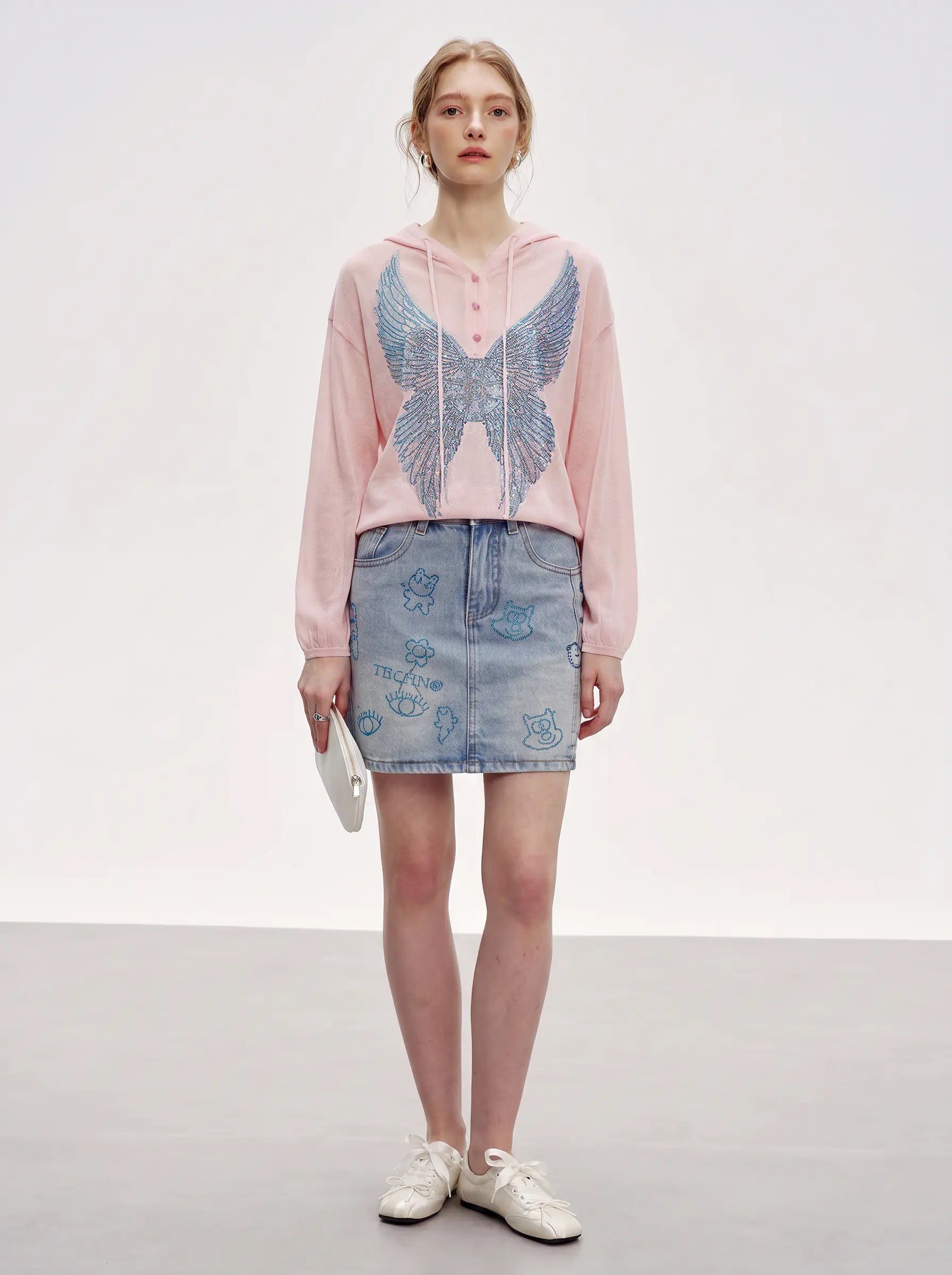A model stands against a light gray studio background wearing a pink cropped hoodie with a large blue embroidered wing design on the front, a light blue denim mini skirt with embroidered details, and white sneakers. TCH Blue Rhinestone Angel Wings Cropped Hoodie